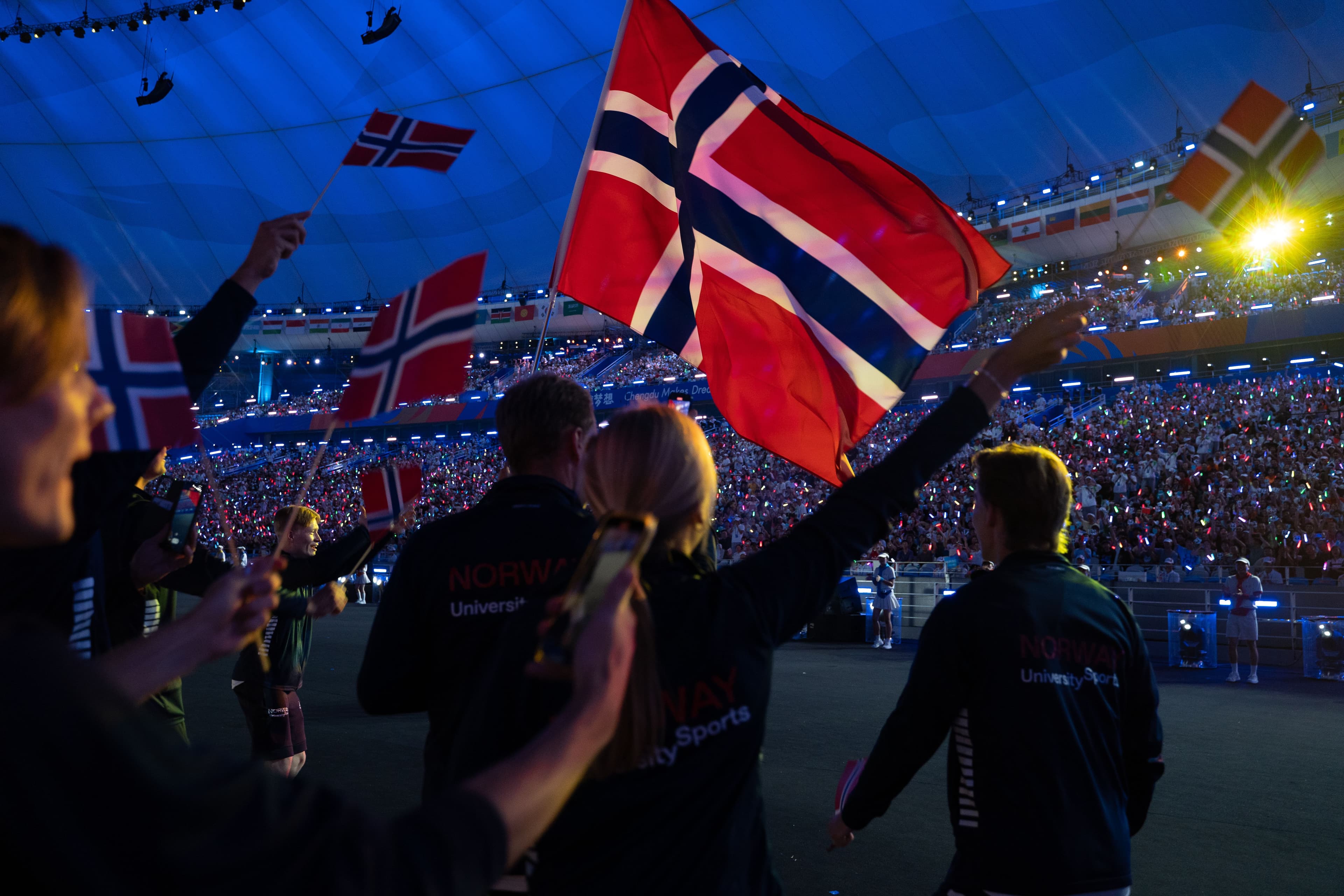 Deltakere vifter med norske flagg under en åpningsseremoni på et fullsatt stadion.
