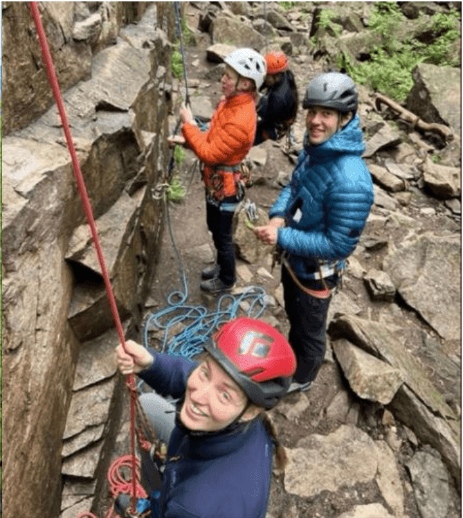 Fire studenter med klatrehjelmer smiler mot kamera under klatring i en bratt fjellvegg.