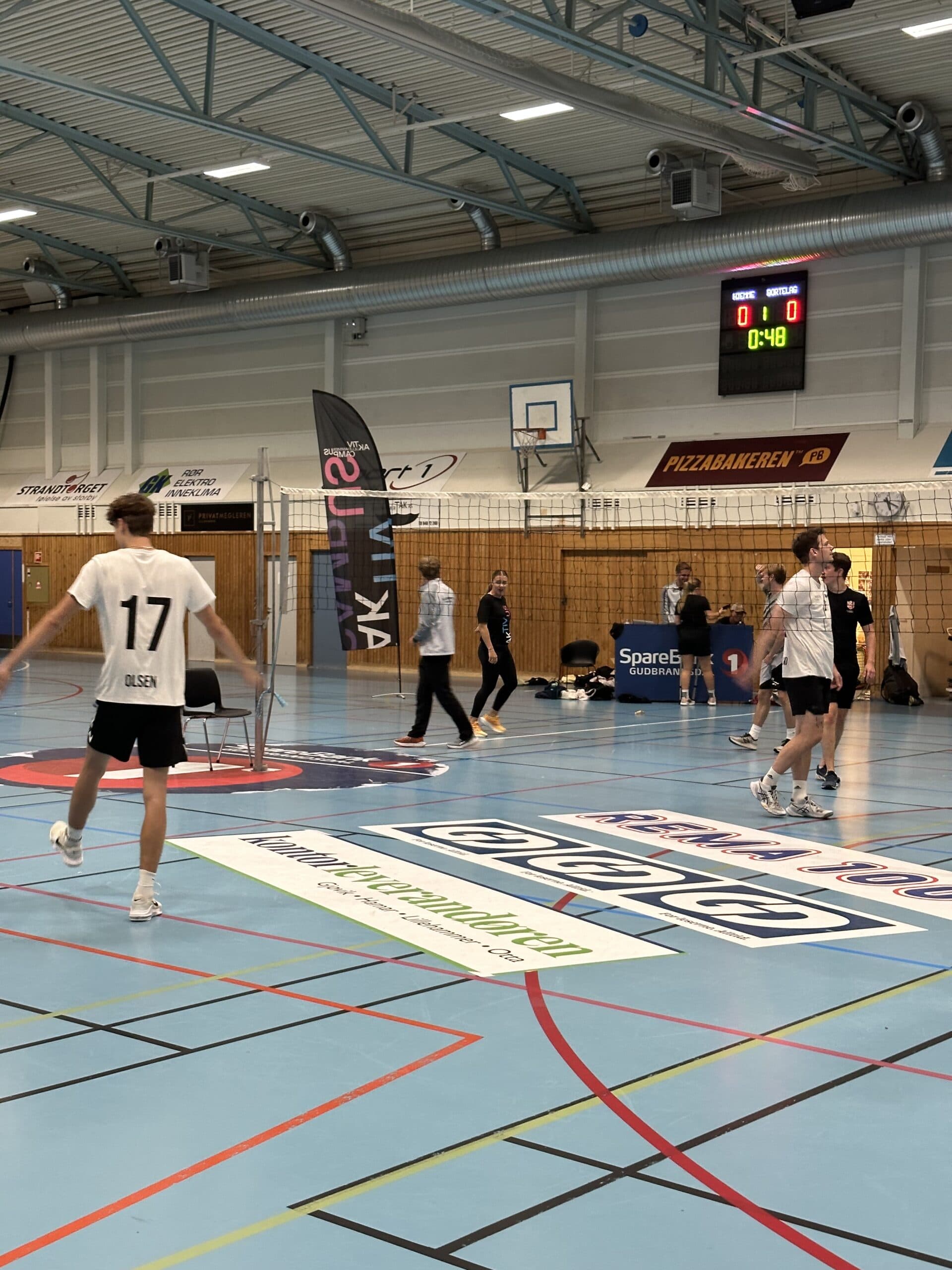 Personer spiller volleyball i en idrettshall med resultattavle og reklamebannere.