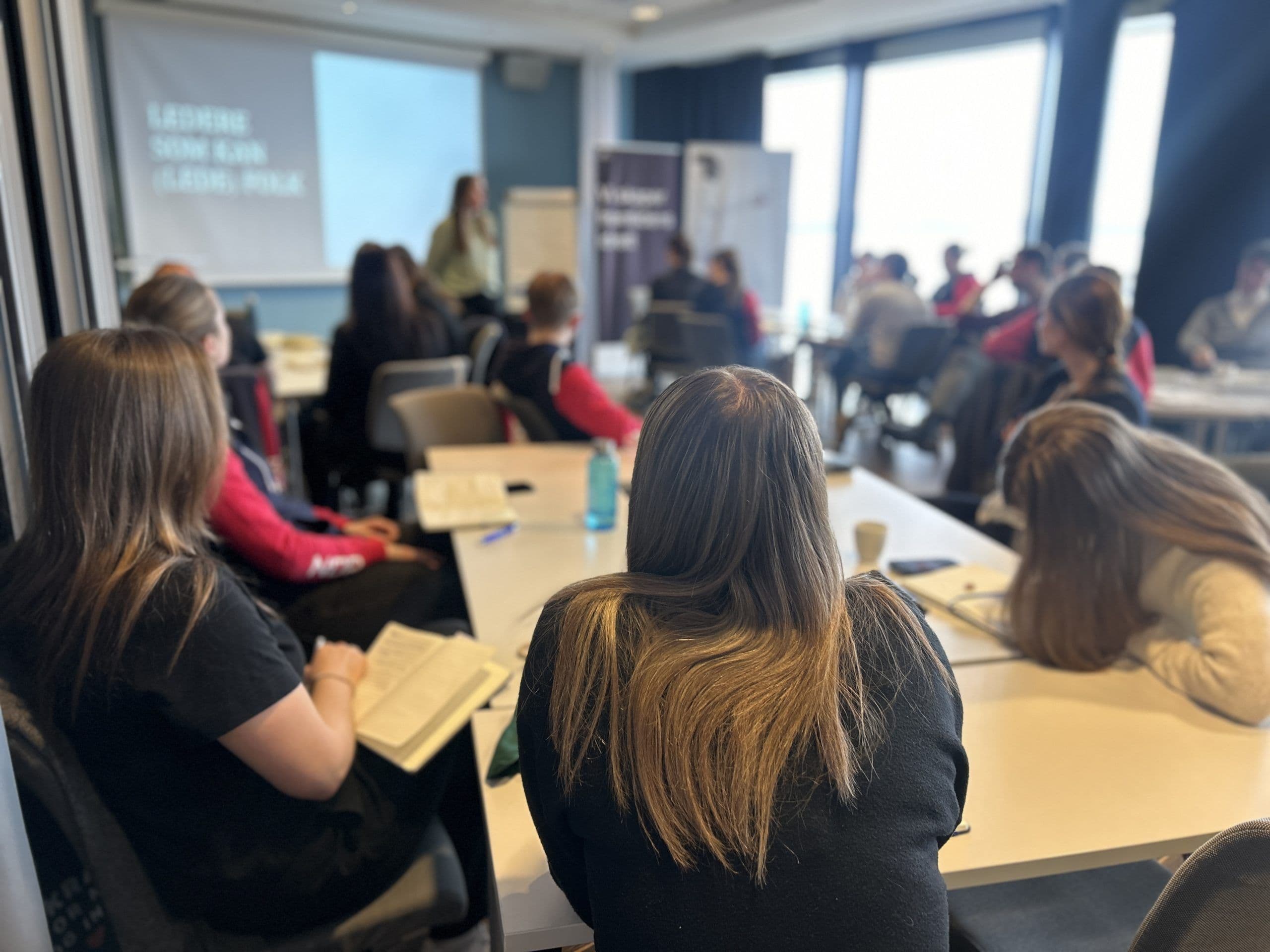 Deltakere sitter ved bord og følger en presentasjon på et seminar.