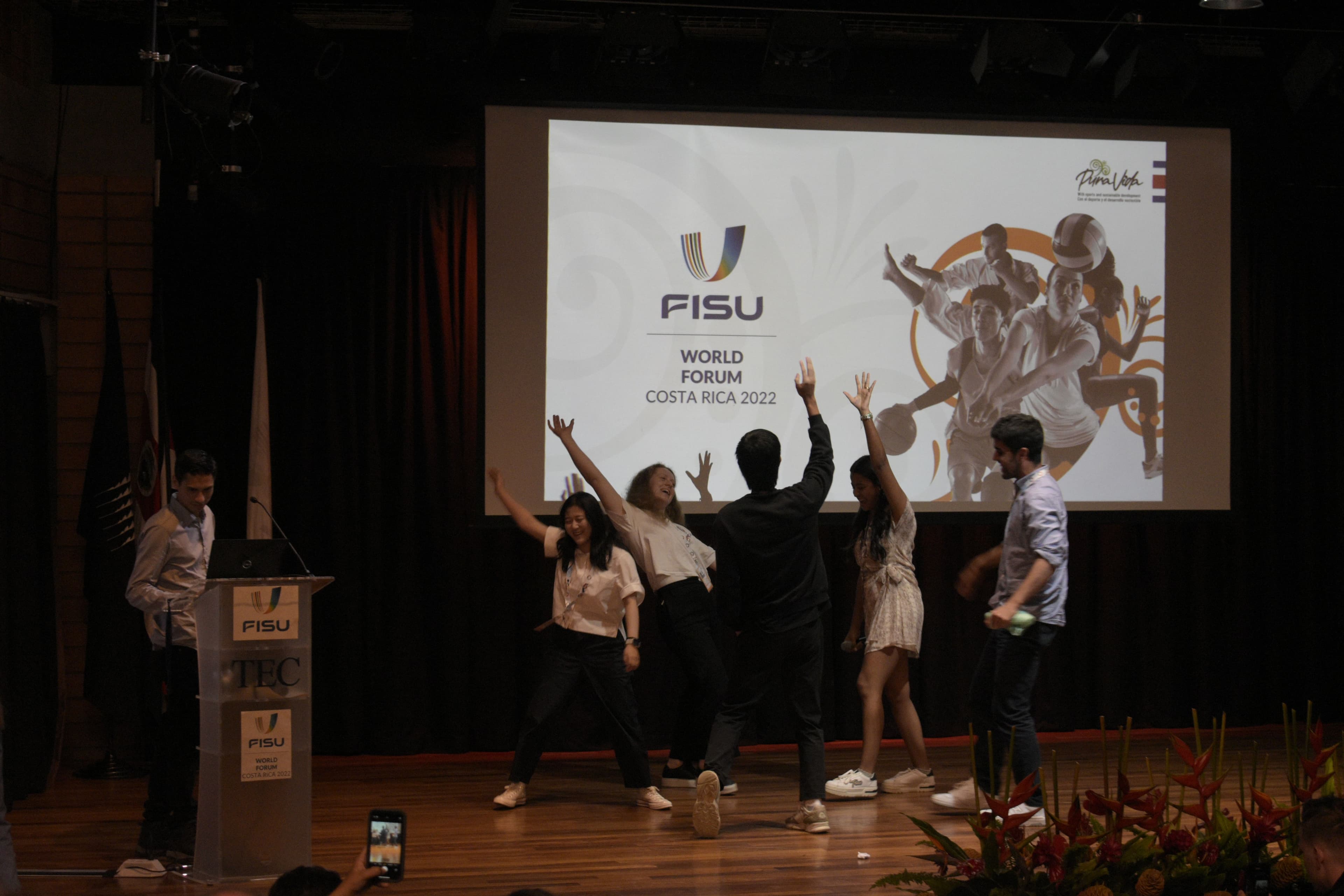 Unge mennesker danser på en scene foran en skjerm med teksten "FISU World Forum Costa Rica 2022".