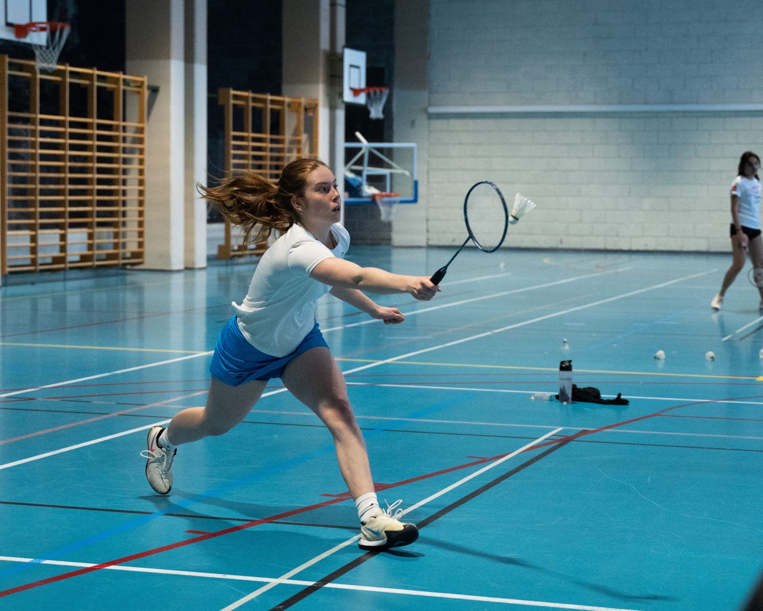 En kvinnelig badmintonspiller strekker seg etter fjærballen med racketen i en idrettshall.