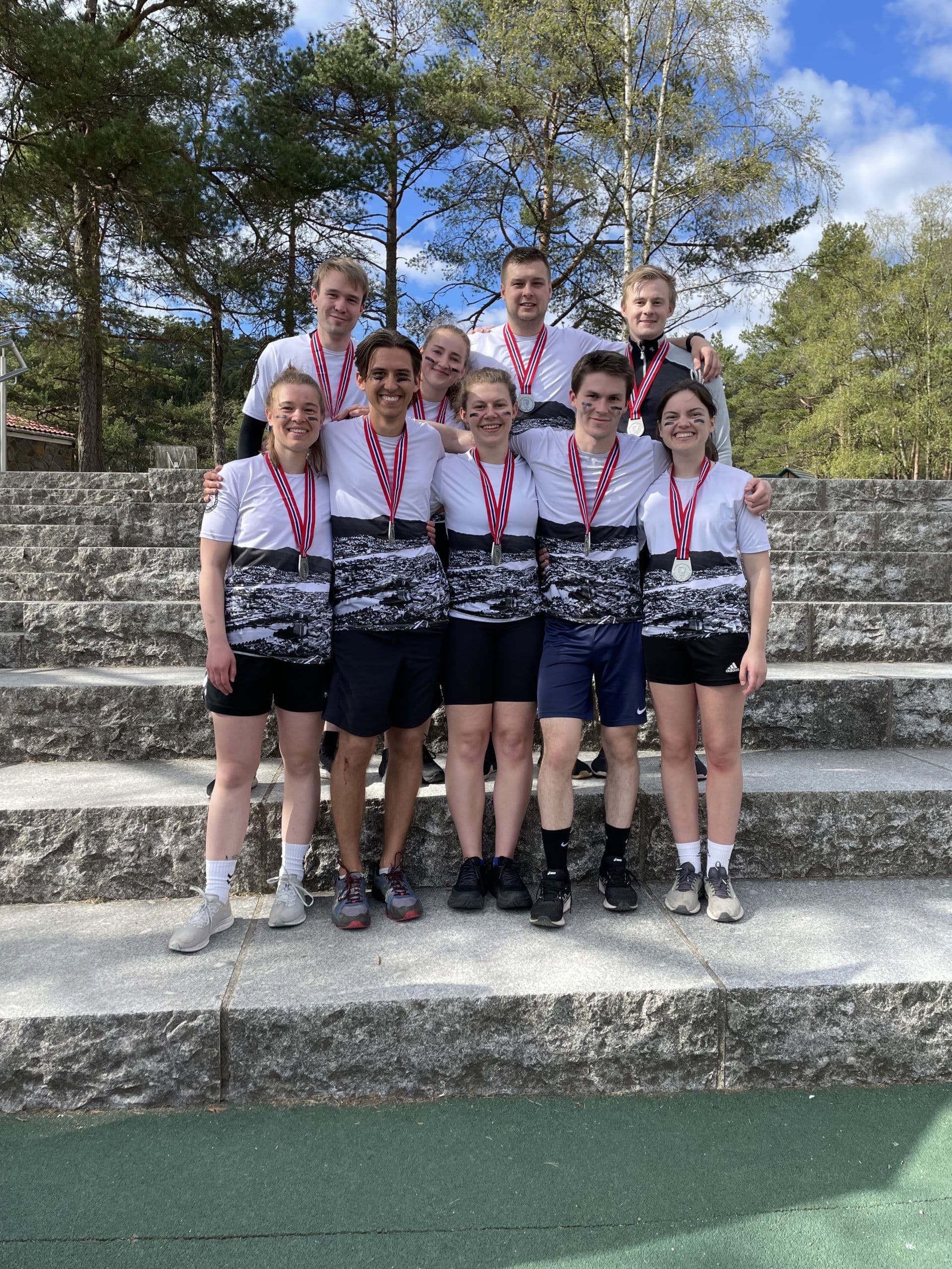 Ni studenter med medaljer rundt halsen smiler samlet på en steintrapp utendørs.