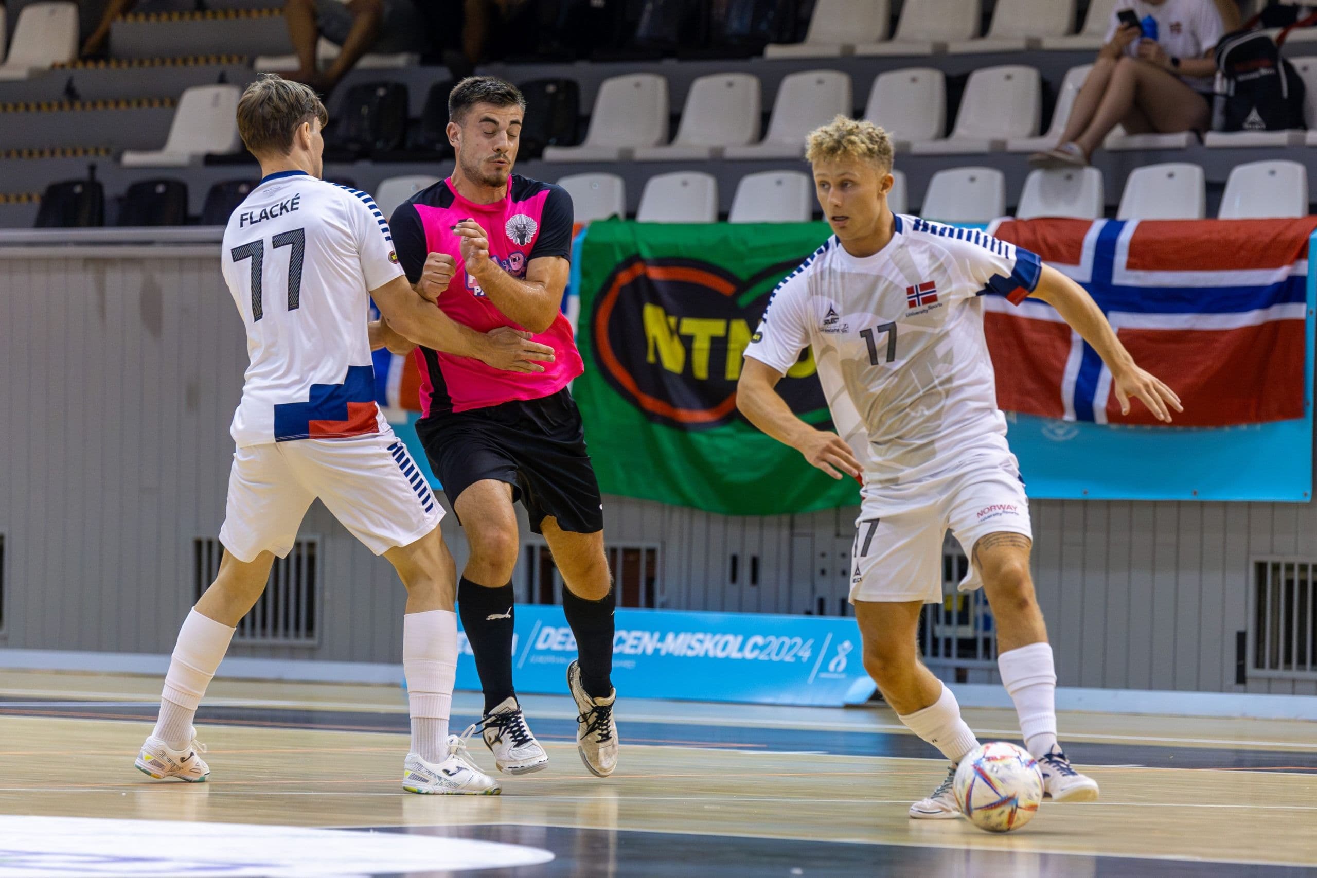 Futsalkamp der to spillere kjemper om ballen foran en dommer, med norsk flagg i bakgrunnen.