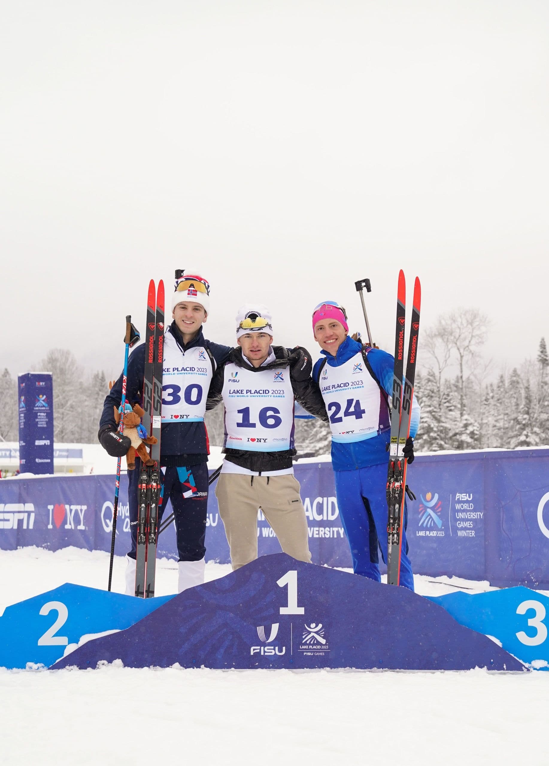 Tre langrennsutøvere med ski poserer på seierspallen under FISU-lekene i Lake Placid 2023.