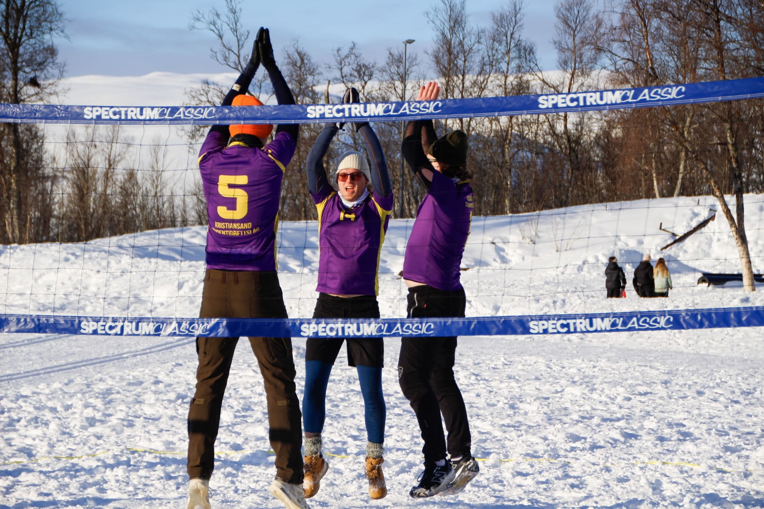 Tre spillere hopper opp mot nettet i en volleyballkamp på snø ute i fjellet.
