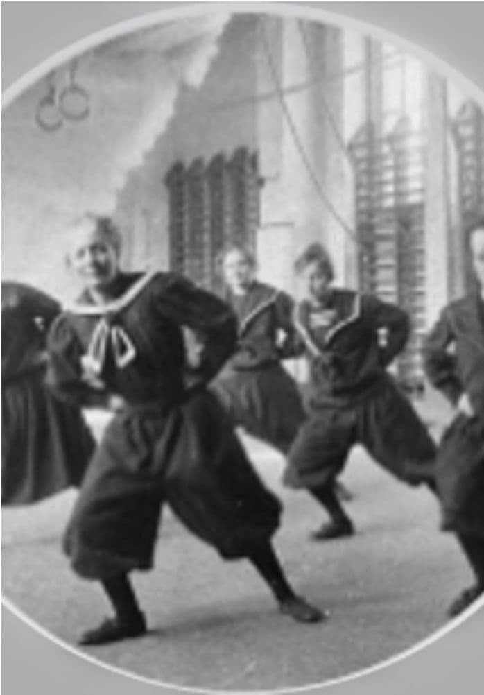 Historisk svart-hvitt-bilde av kvinner i matrosdrakter som utfører gymnastikk i en sal.