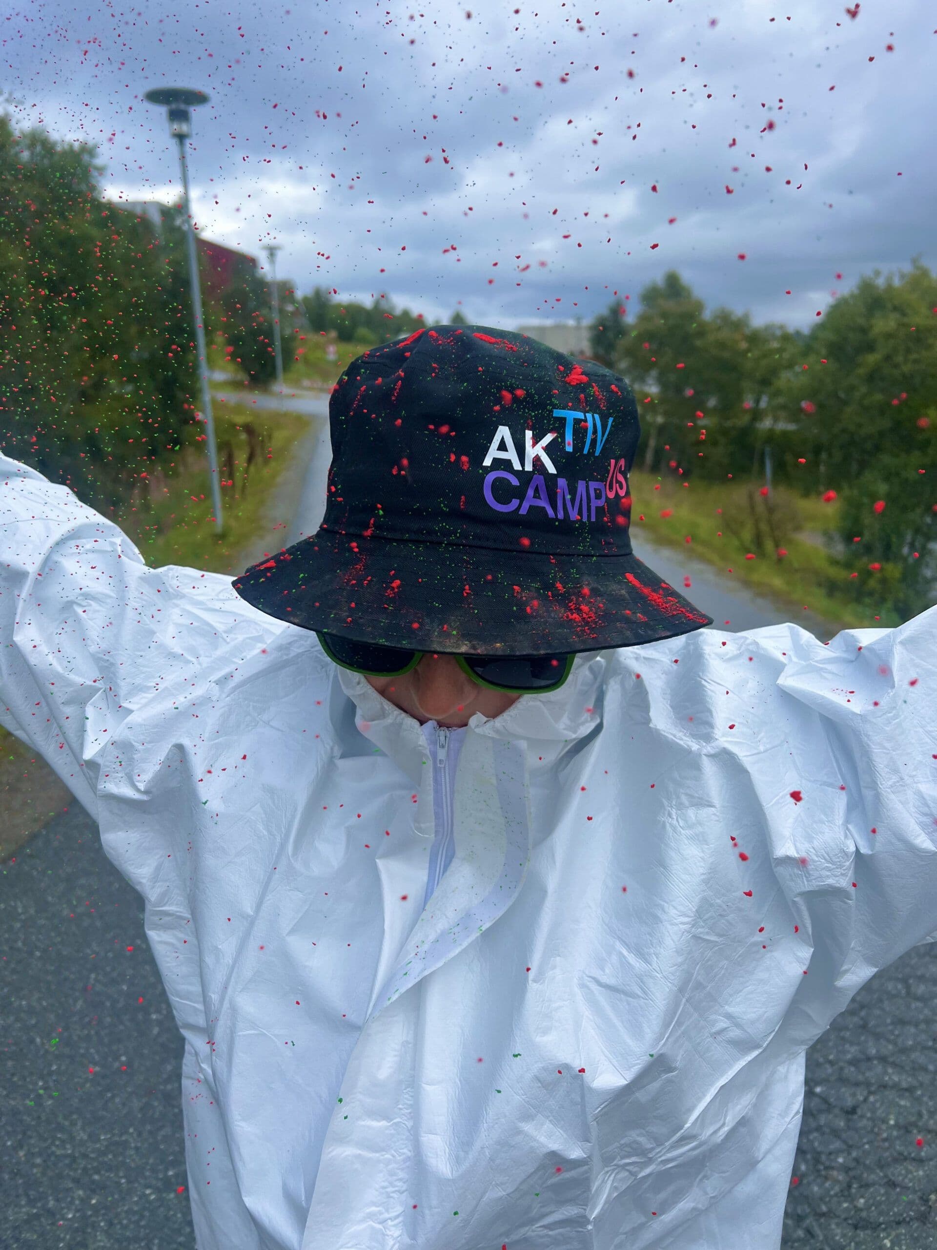 En person i hvit vernedress og bøttehatt med teksten «Aktiv Campus», dekket av røde fargeflekker utendørs.