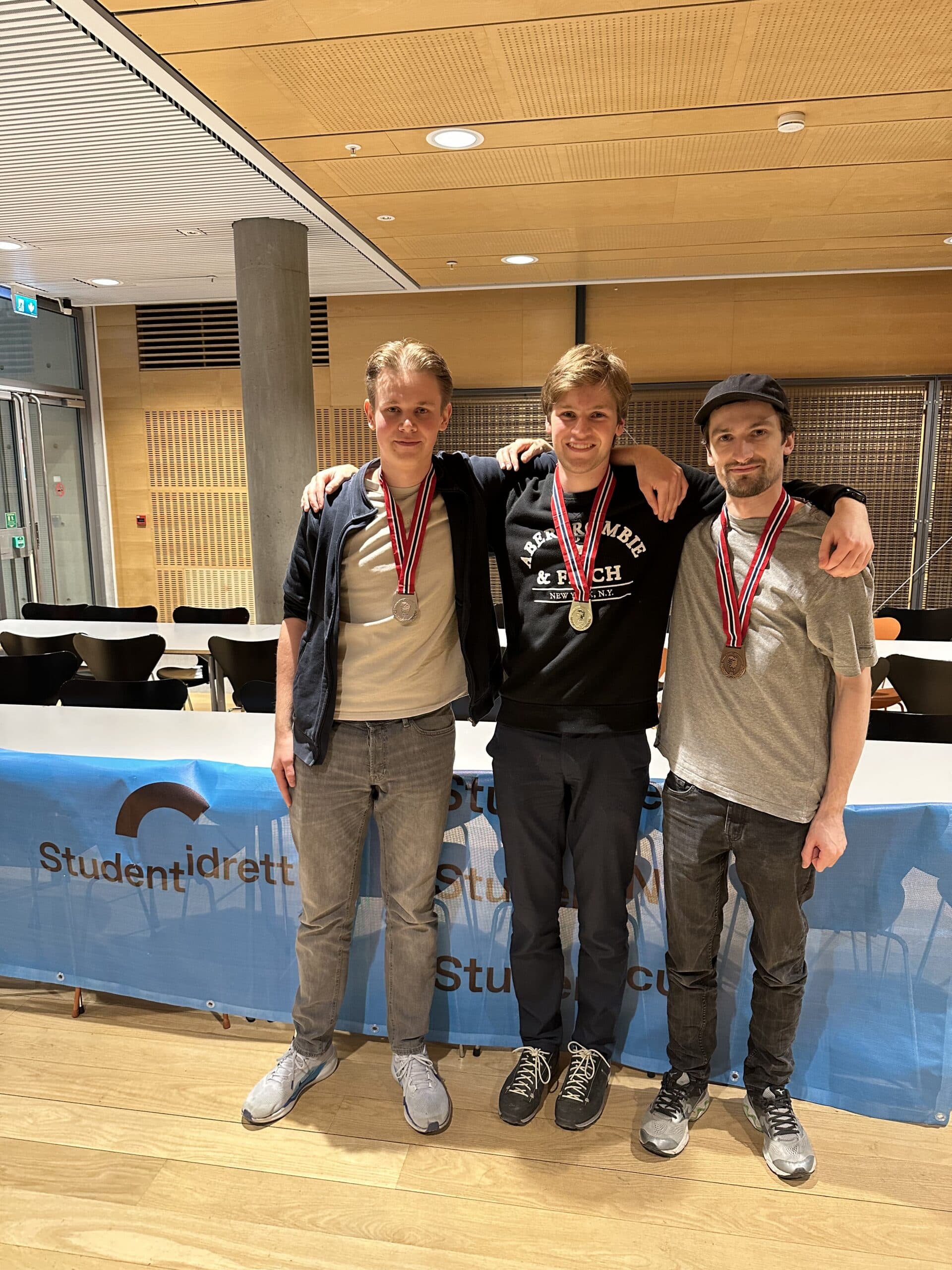 Tre menn med medaljer rundt halsen står med armene rundt hverandre foran en Studentidrett-banner.
