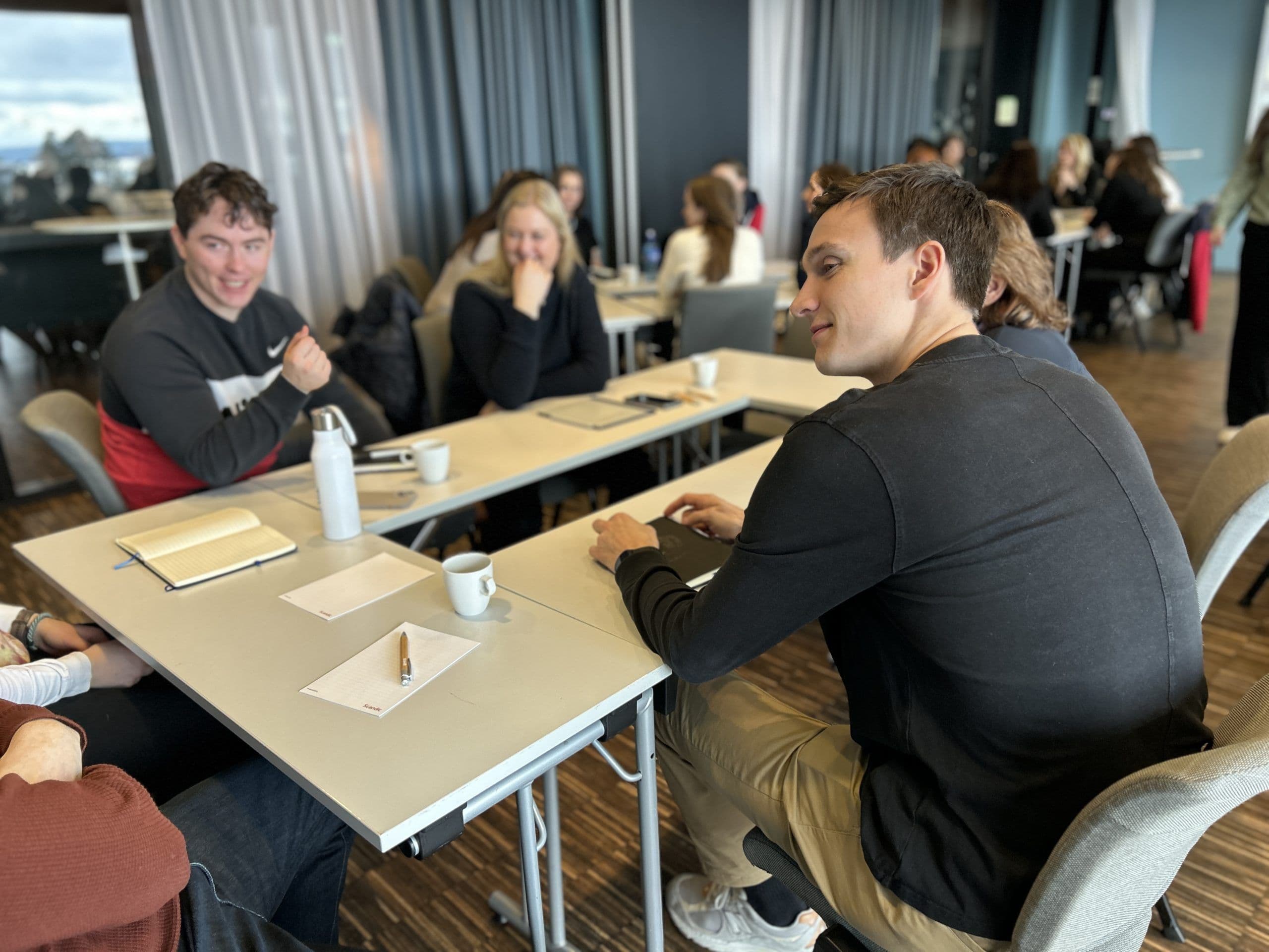 Studenter sitter rundt bord og samtaler under en samling med utsikt over vann.