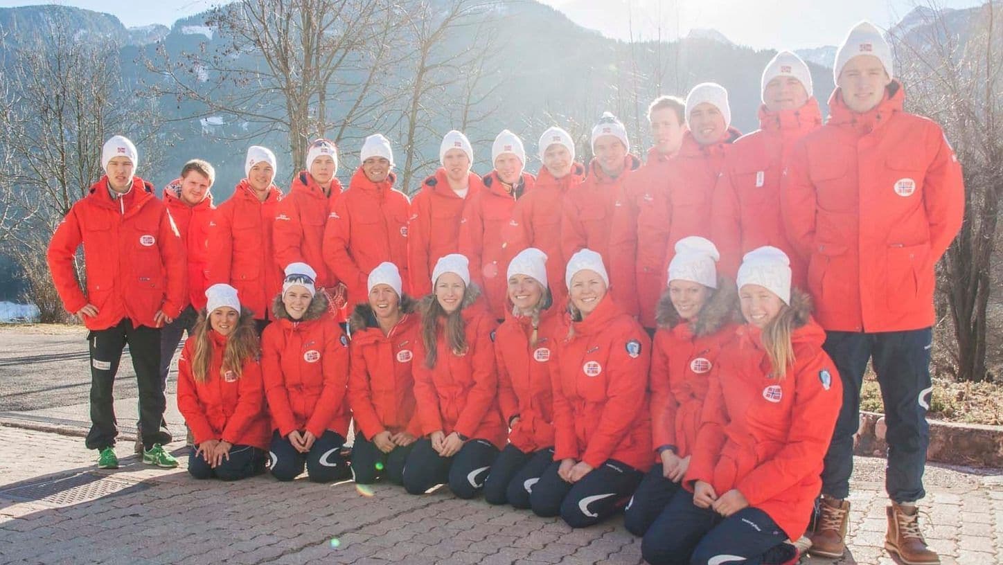 En gruppe utøvere i røde og hvite landslagsjakker og luer poserer utendørs foran et fjellandskap om vinteren.