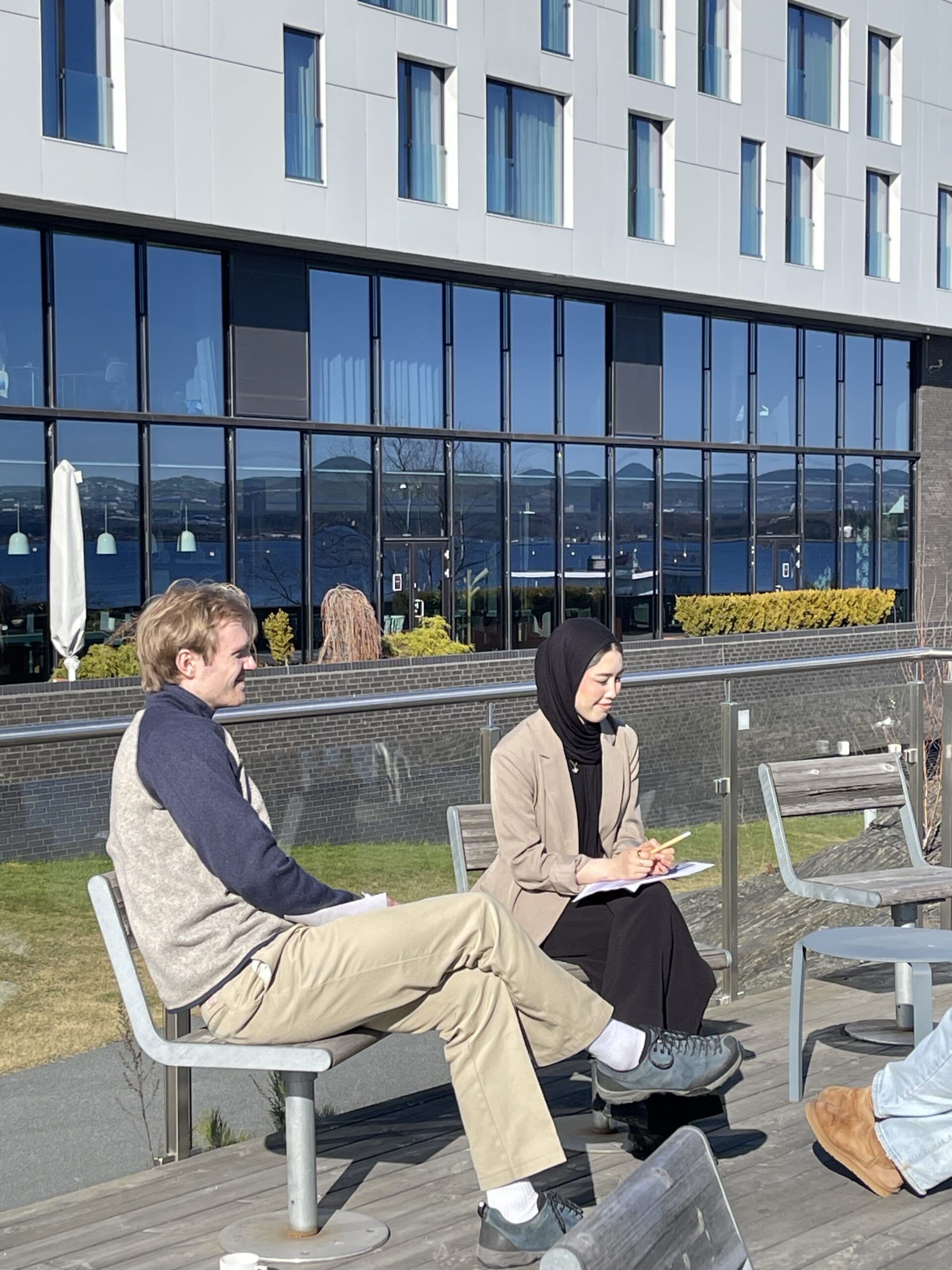 En mann og en kvinne sitter og samtaler på en uteplass med notatblokker foran en bygning ved vannet.