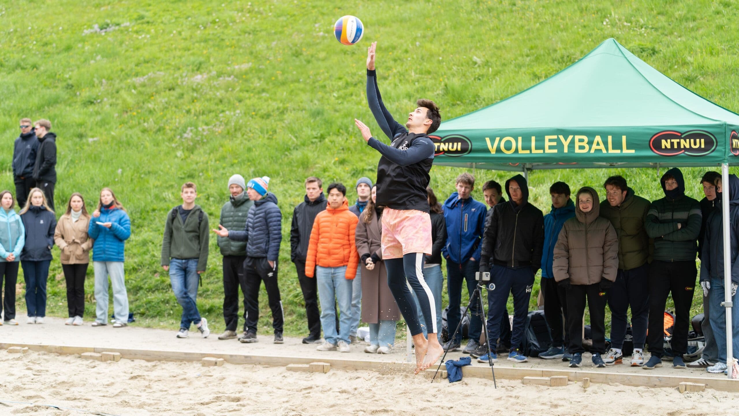 En volleyballspiller server ballen på en sandbane mens et publikum følger med.