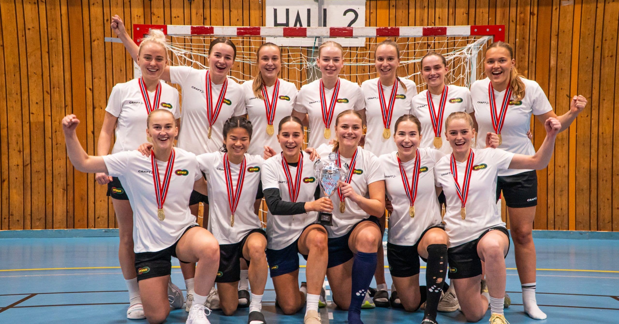 Et jublende jentelag i hvite drakter poserer med pokal og gullmedaljer i en idrettshall.