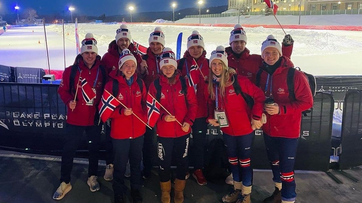 En gruppe utøvere i røde landslagsklær poserer med norske flagg foran en olympisk arena om kvelden.