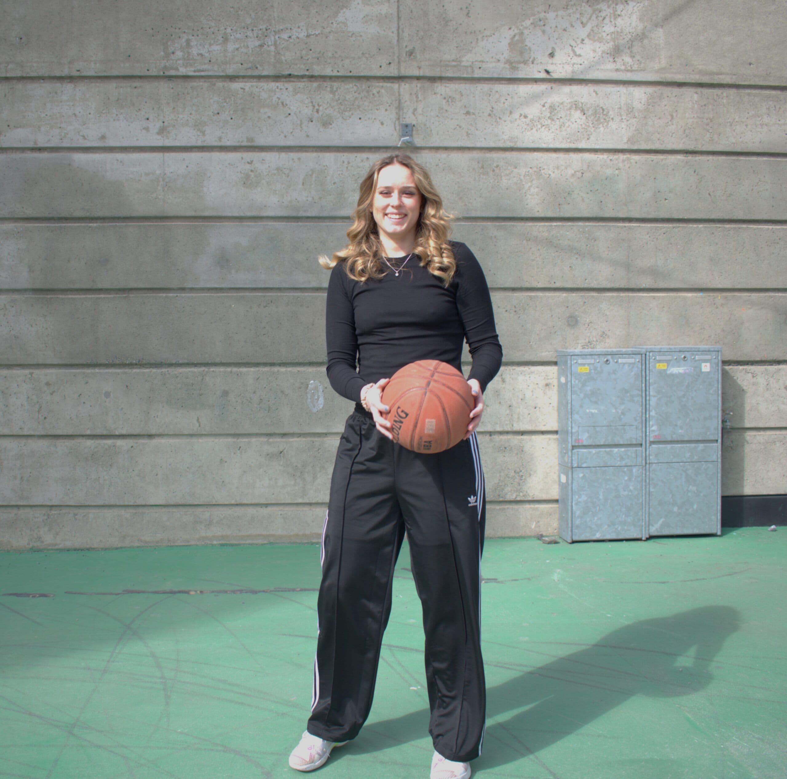 En smilende kvinne holder en basketball og står foran en betongvegg på en utebane.