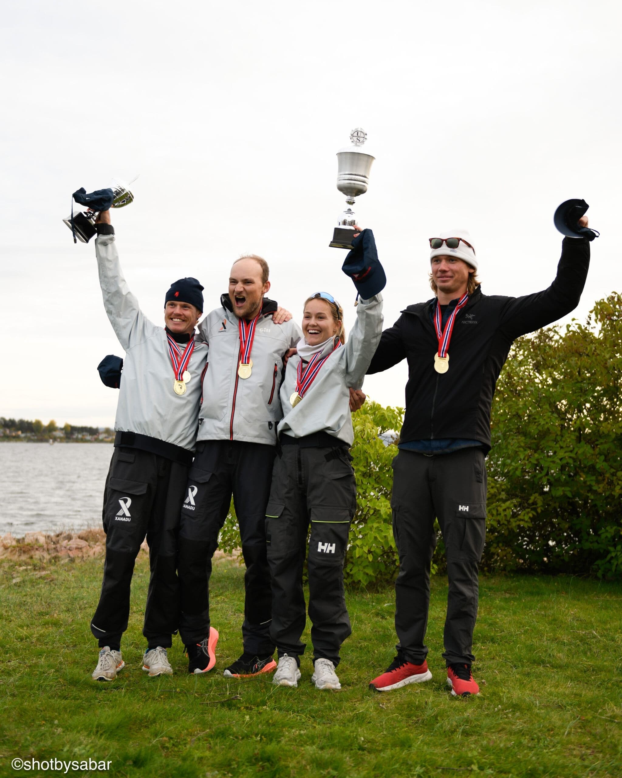 Fire seilere med medaljer jubler med pokal og luer i været utendørs ved vannet.