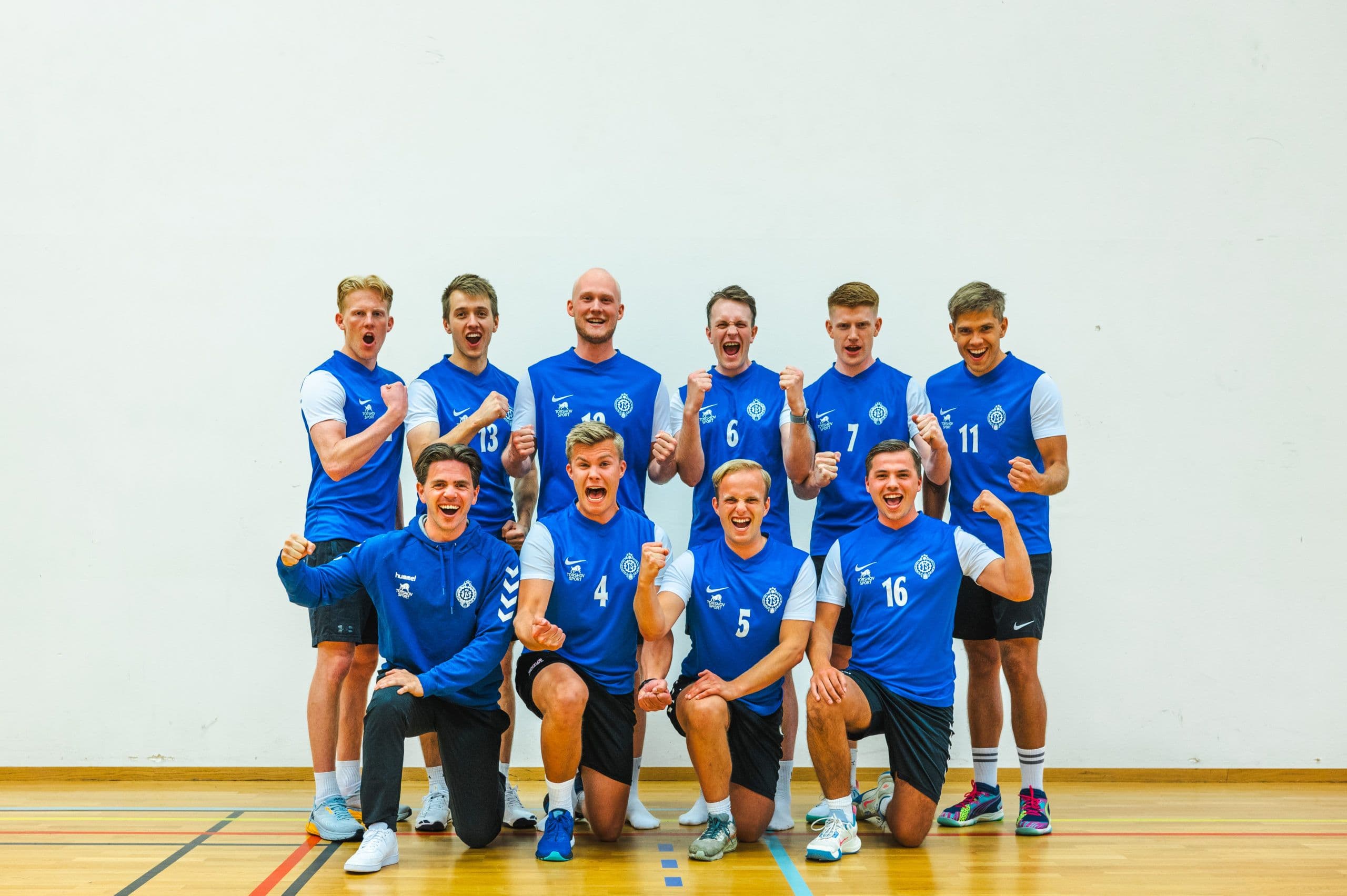 Et herrelag i volleyball poserer samlet i blå drakter i en idrettshall.