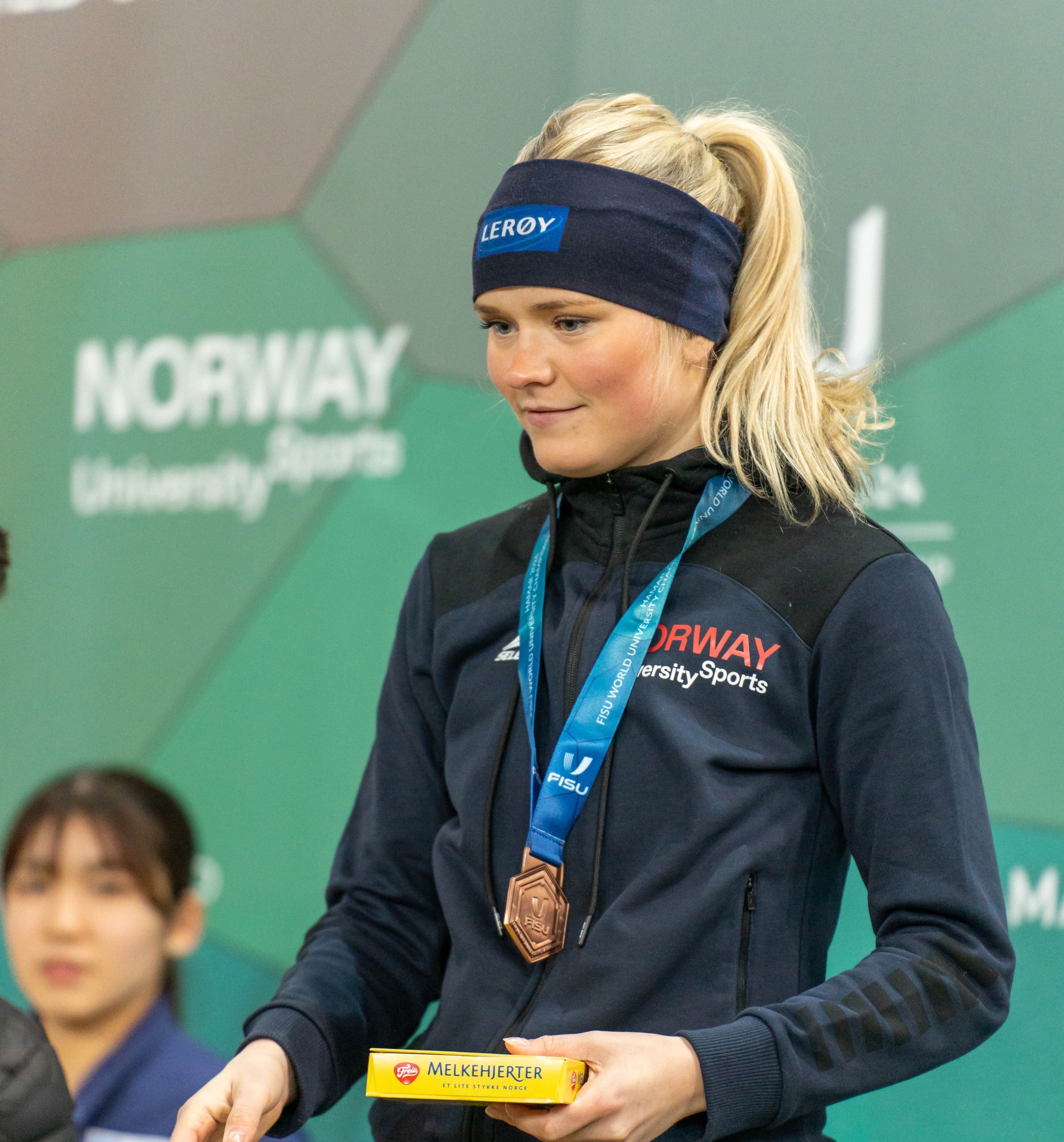 En kvinnelig utøver står på seierspallen med bronsemedalje og jakke fra Norway University Sports.