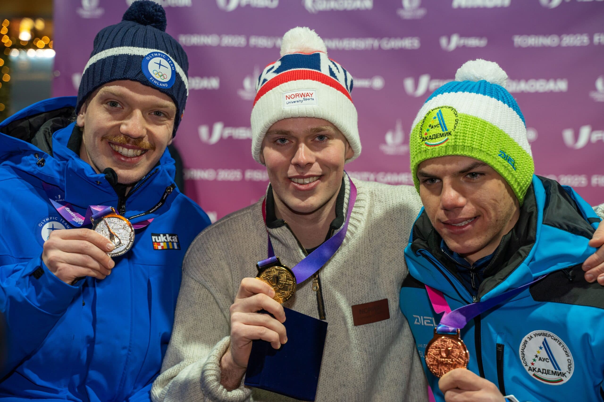 Tre mannlige utøvere med luer viser fram medaljene sine foran en vegg med teksten Torino 2025 FISU University Games.