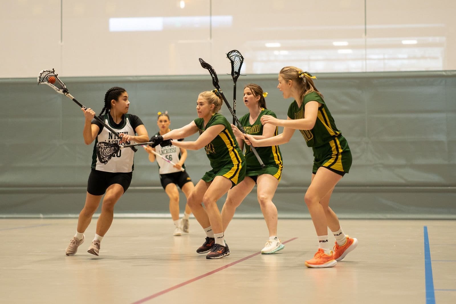 Kvinnelige spillere i lacrosse kjemper om ballen med køller i en idrettshall.