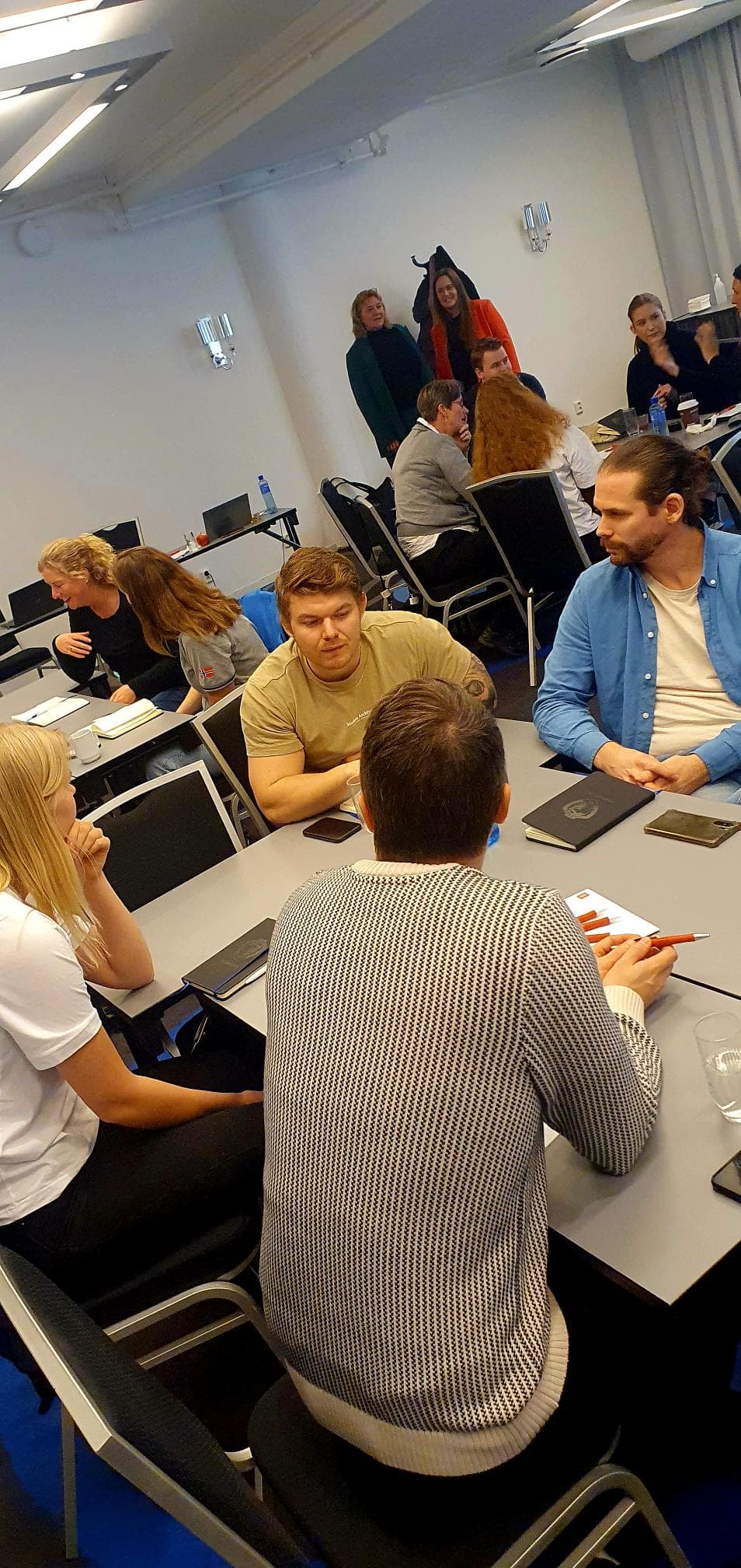 Studenter sitter og samtaler rundt bord i et møterom eller seminarlokale.