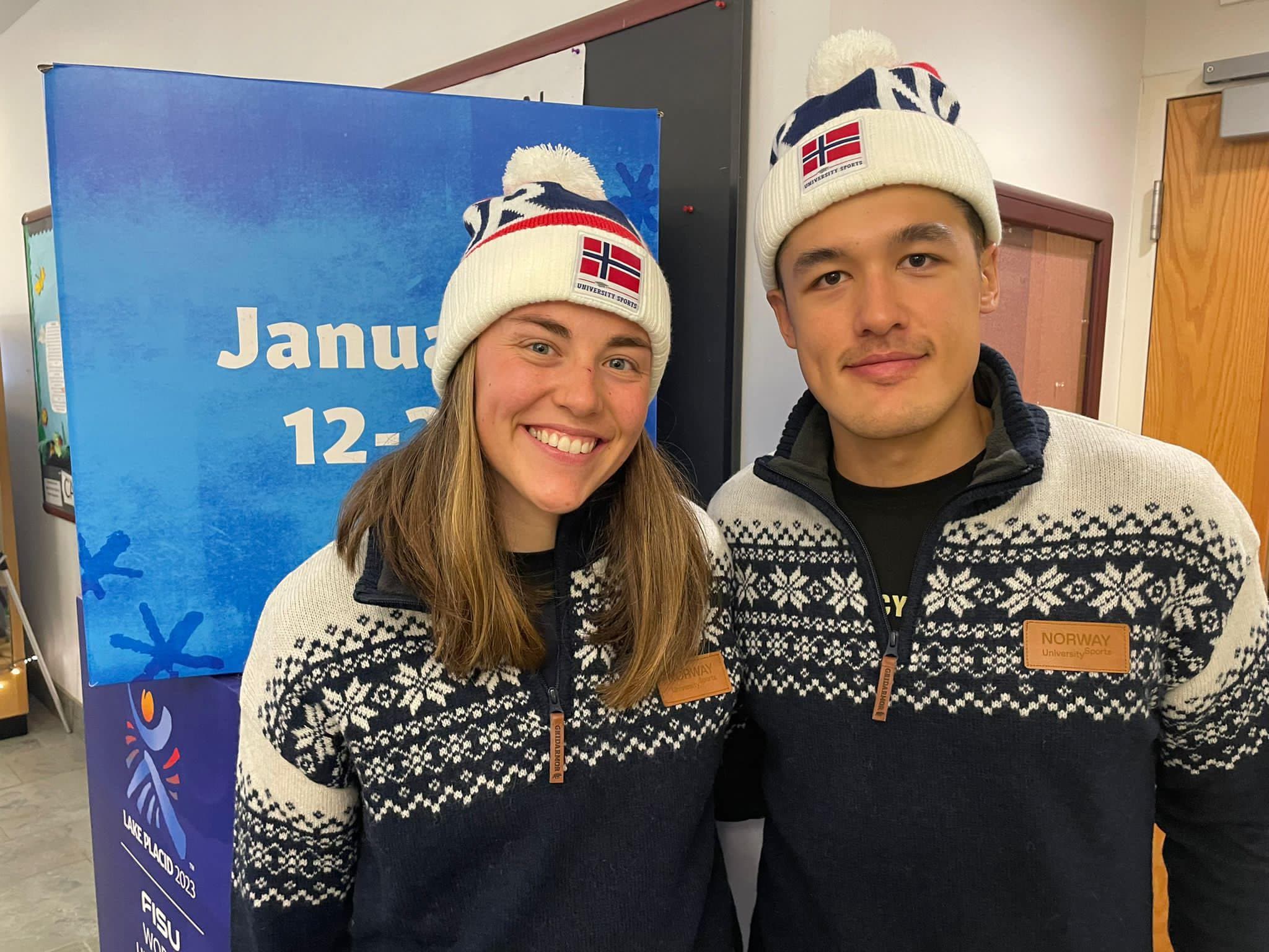 En ung kvinne og en ung mann i Norge-vinterantrekk og luer smiler foran en FISU-plakat.
