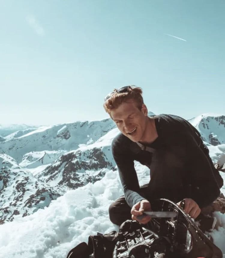 En ung mann smiler på en snødekt fjelltopp med snødekte fjell i bakgrunnen.