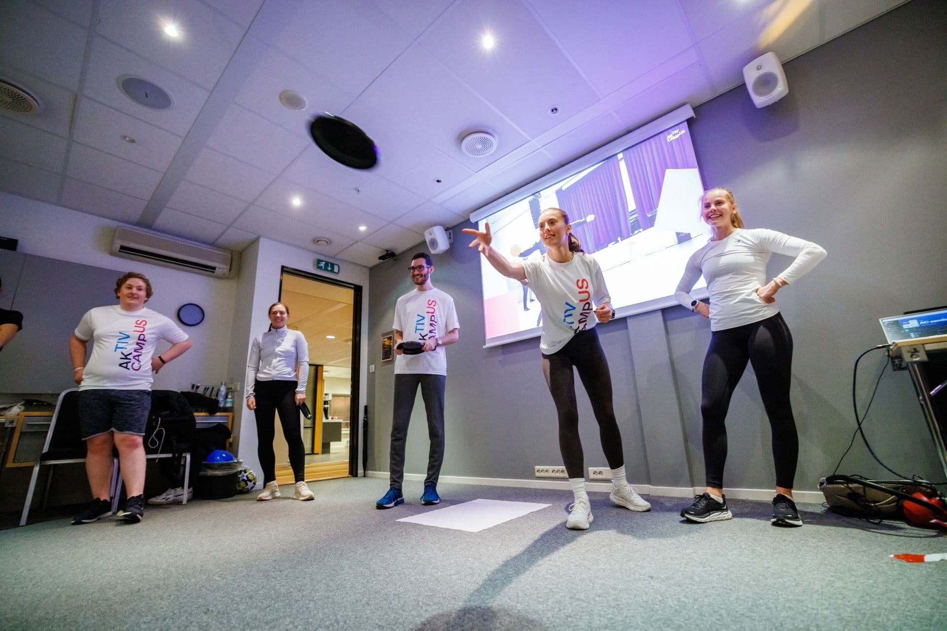 En gruppe studenter i Aktiv på campus-trøyer gjør øvelser i et rom med projektor.