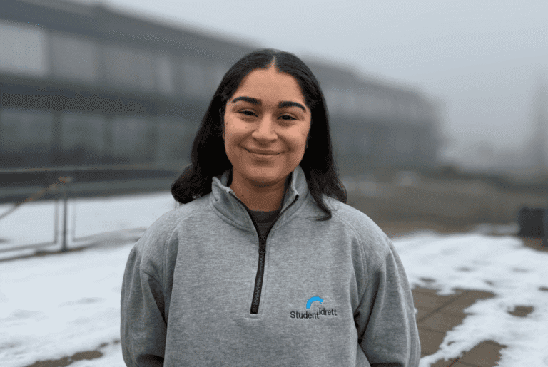 Portrett av en smilende ung kvinne i grå Studentidrett-genser utendørs.
