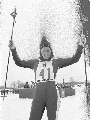 Historisk svart-hvitt-foto av en kvinnelig langrennsløper med startnummer 41 som løfter stavene i været.