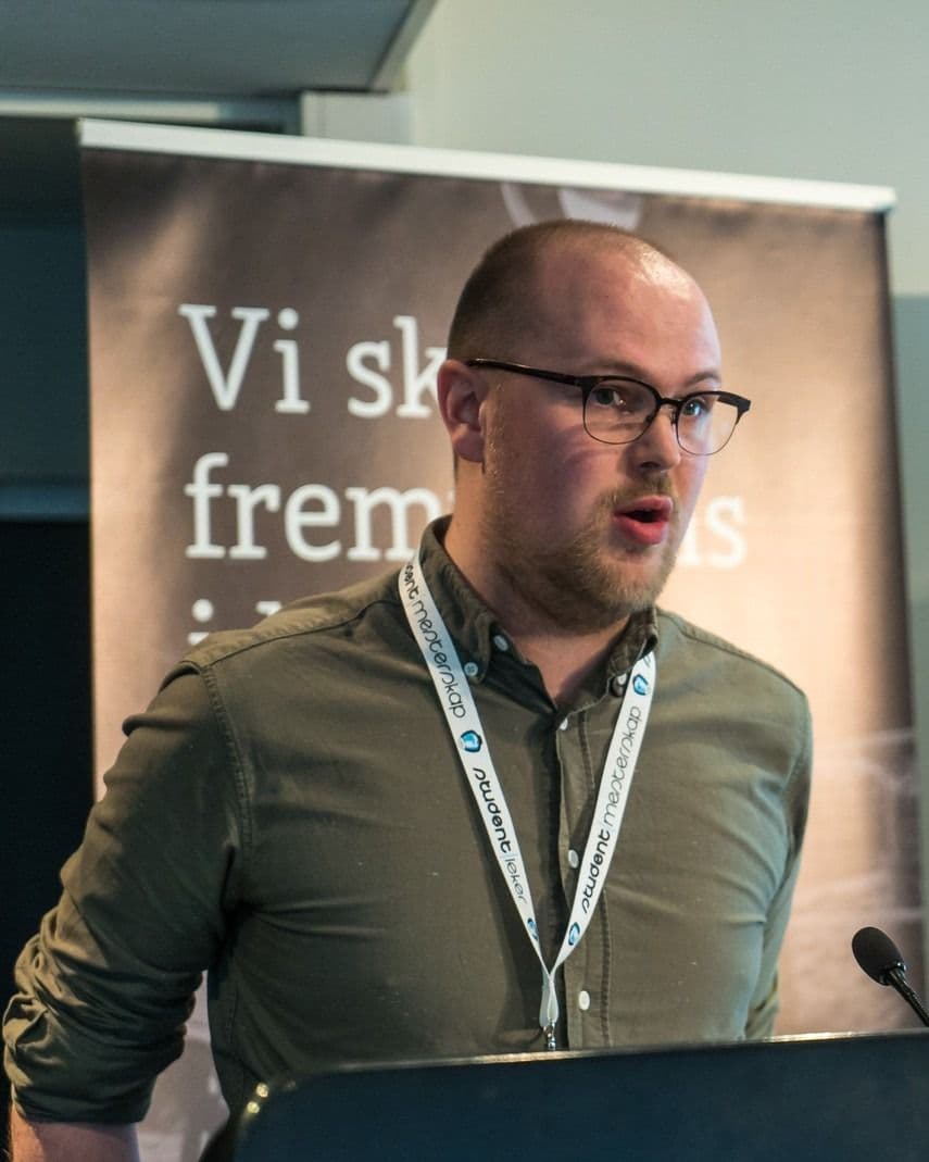 En mann med briller taler fra en talerstol foran en Studentidrett-rollup.