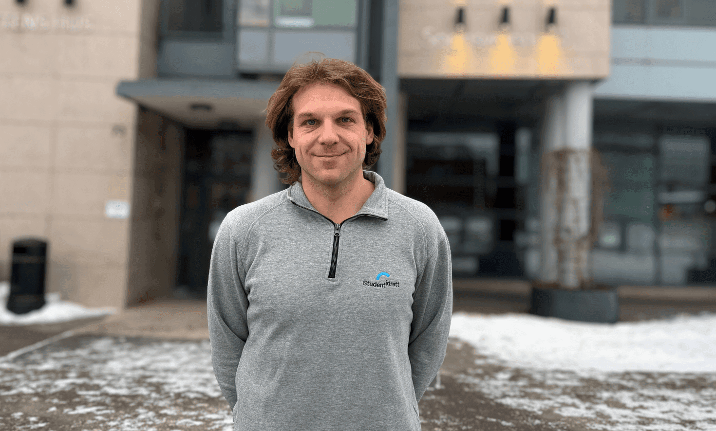 Portrett av en mann i grå Studentidrett-genser foran en bygning.