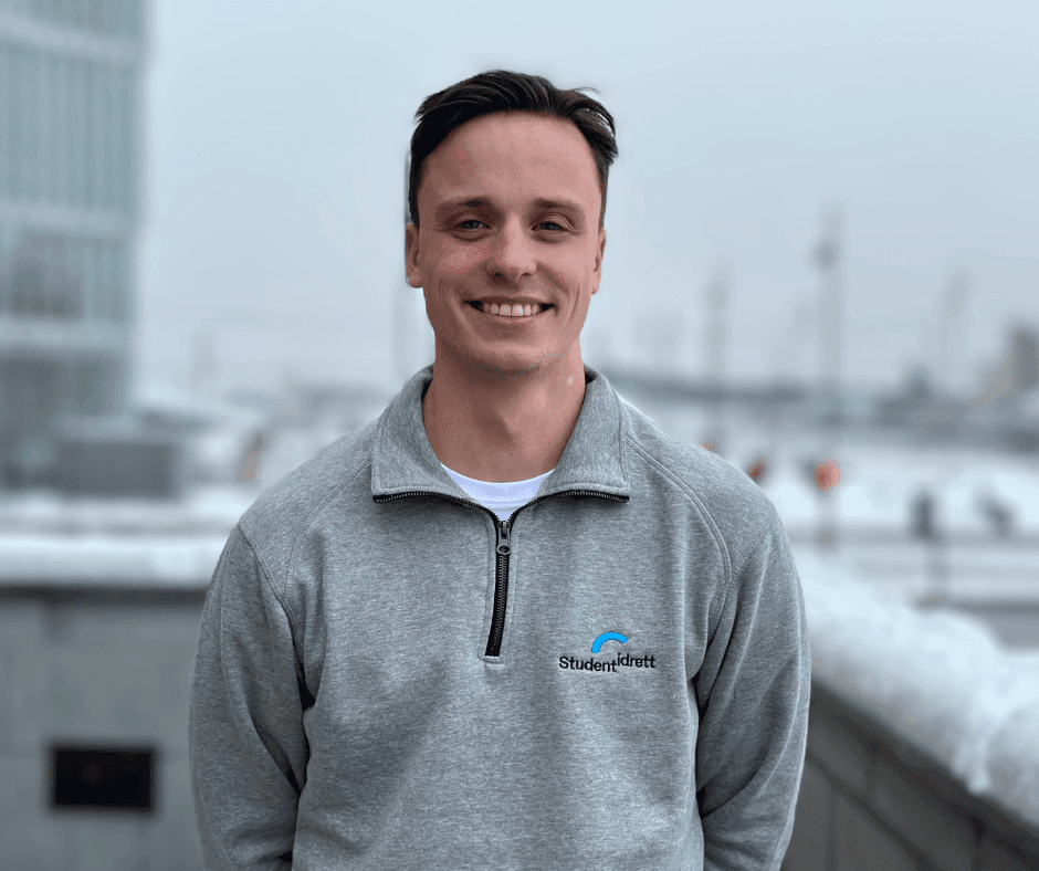 Portrett av en smilende mann i grå Studentidrett-genser med en havn i bakgrunnen.