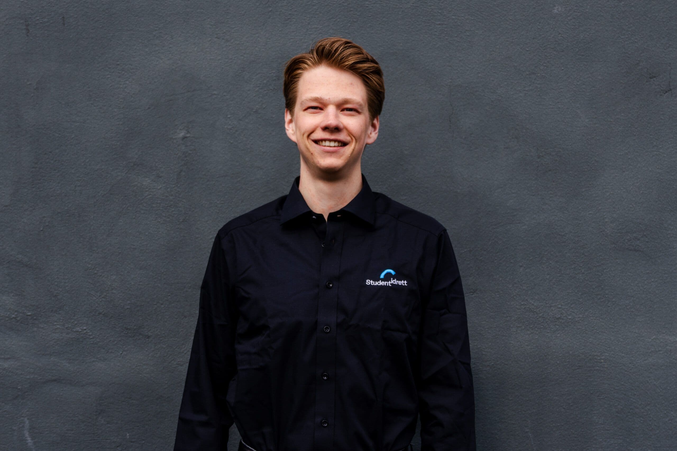 Portrett av en mann i svart Studentidrett-skjorte foran en grå vegg.