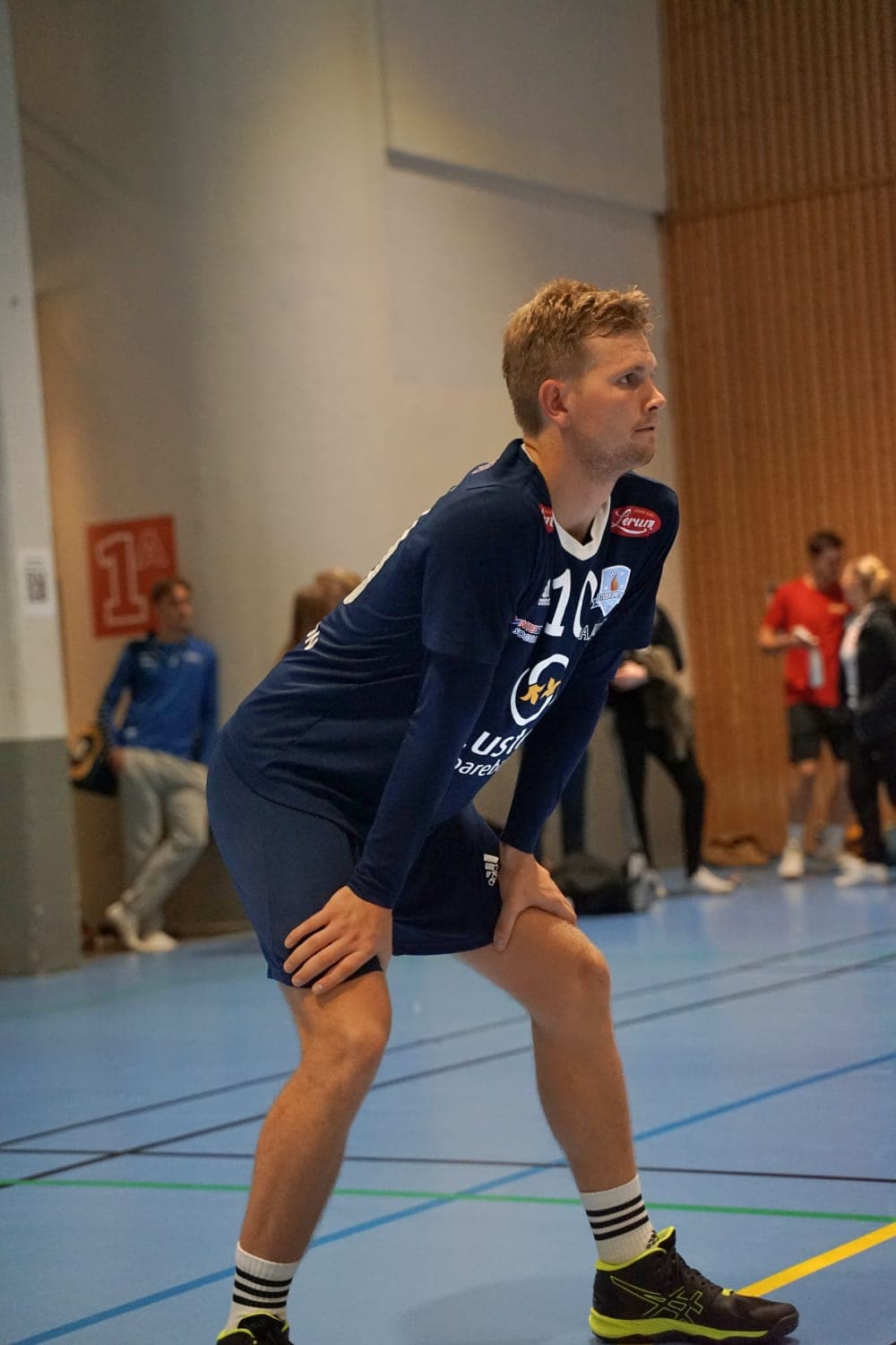 En håndballspiller i mørkeblå drakt står bøyd med hendene på knærne i en idrettshall.