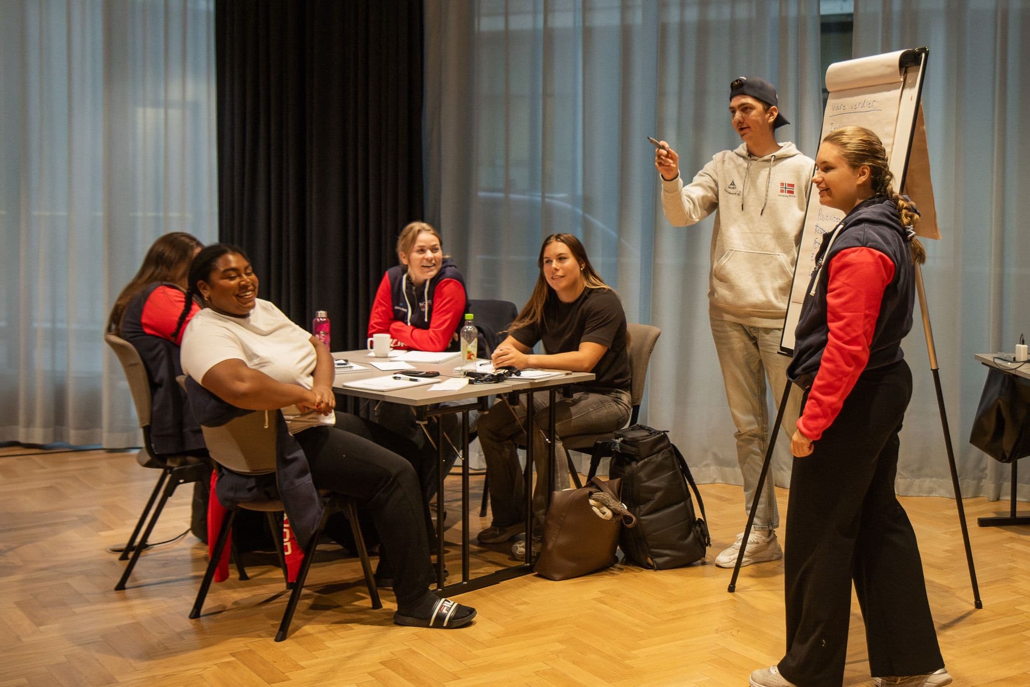 En gruppe studenter deltar i en workshop der en person presenterer ved en flippover.