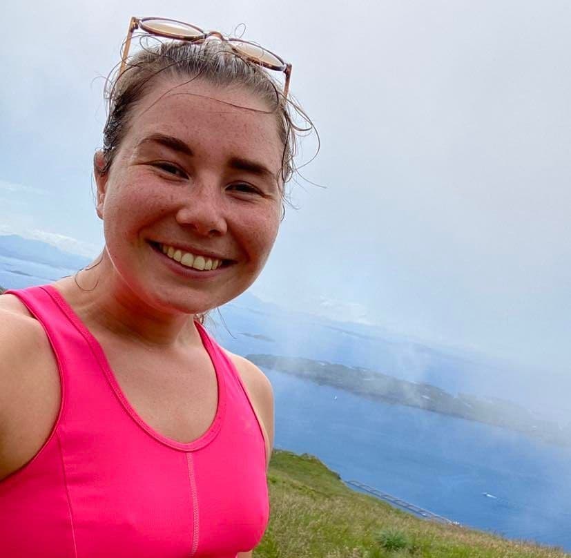 Portrett av en smilende kvinne i rosa treningstopp på en fjelltopp med sjøen i bakgrunnen.