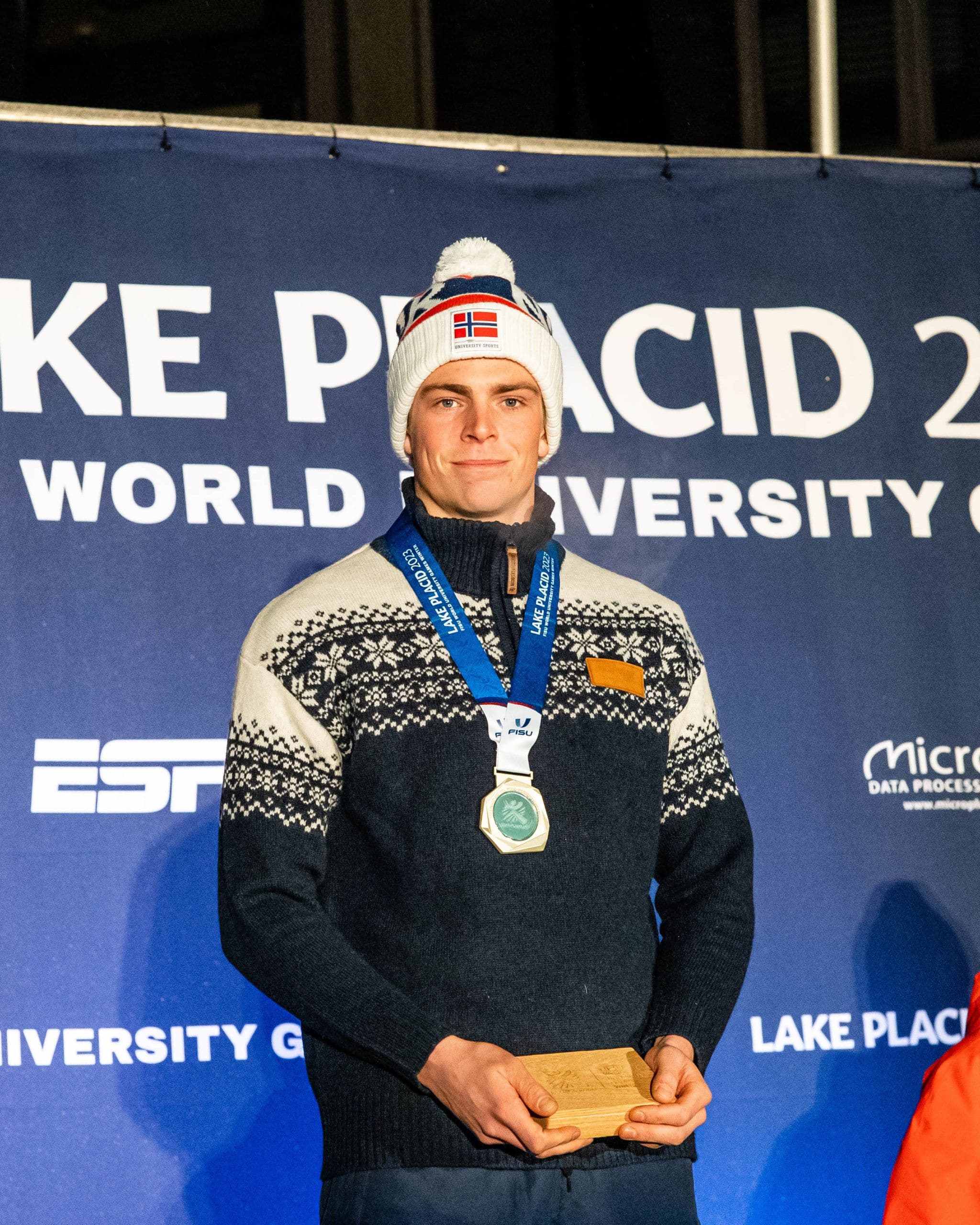 En utøver med sølvmedalje foran en vegg med teksten «Lake Placid 2024 World University Games».