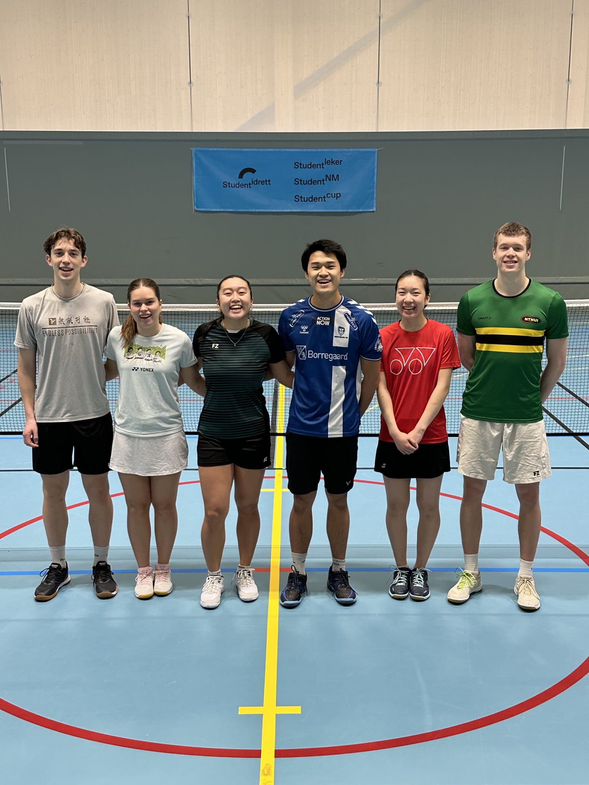 Seks badmintonspillere står på rekke ved nettet i en idrettshall under en Studentidrett-banner.