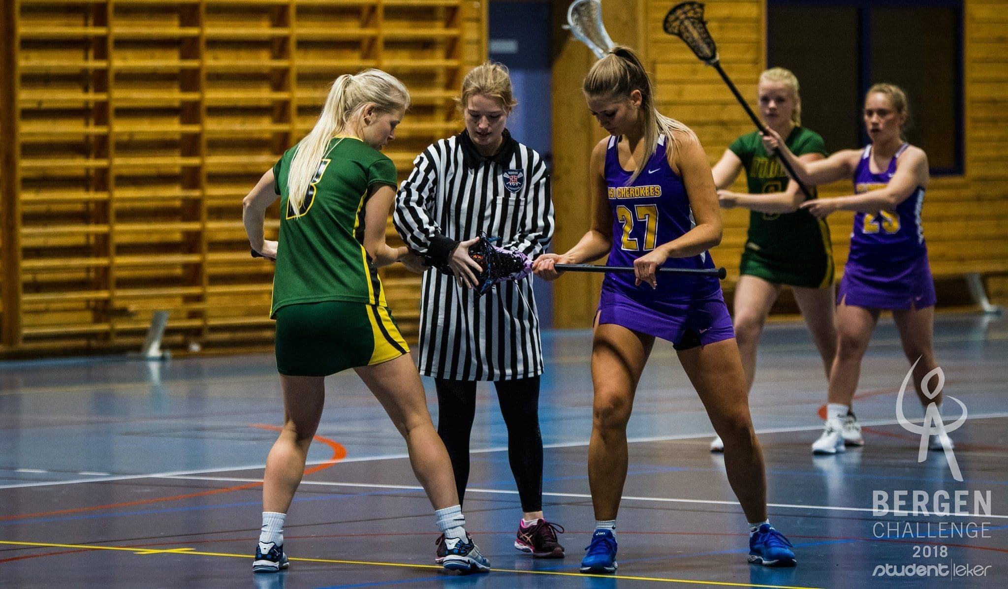 To kvinnelige lacrossespillere og en dommer under en oppkast-situasjon i en idrettshall under Bergen Challenge 2018.