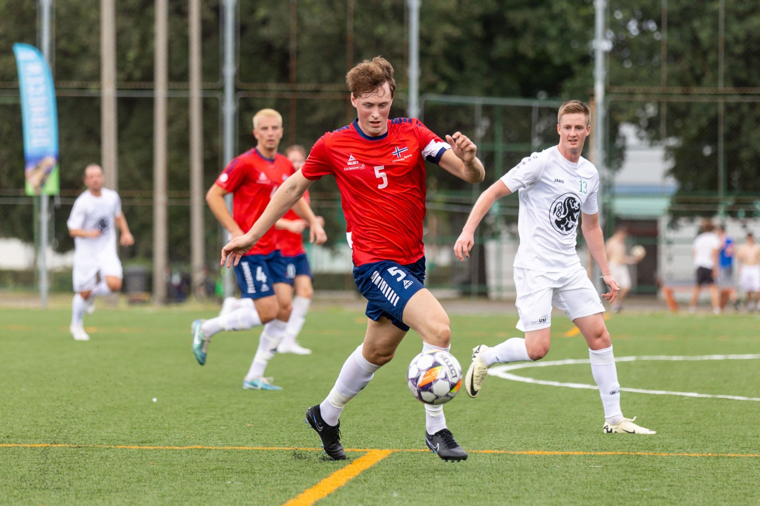 En fotballspiller i rød drakt med nummer 5 fører ballen forbi motspillere i hvitt under en kamp.