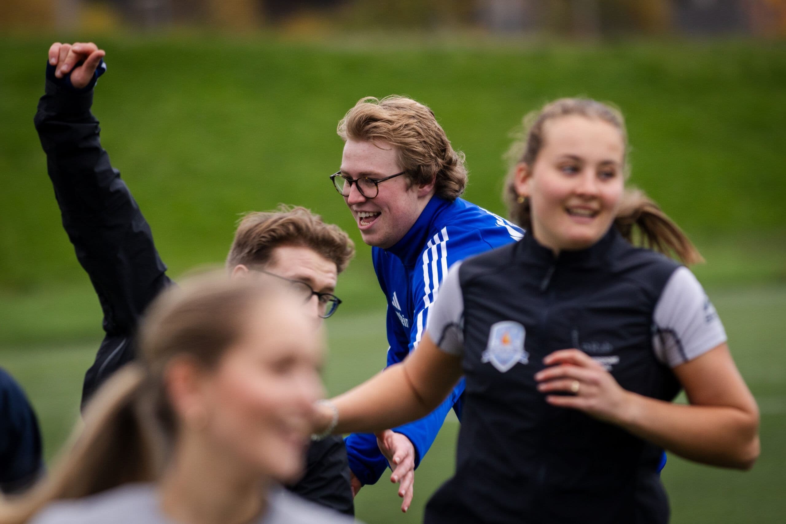 Smilende studenter i aktivitet på en gressbane, en av dem løfter armen i været.