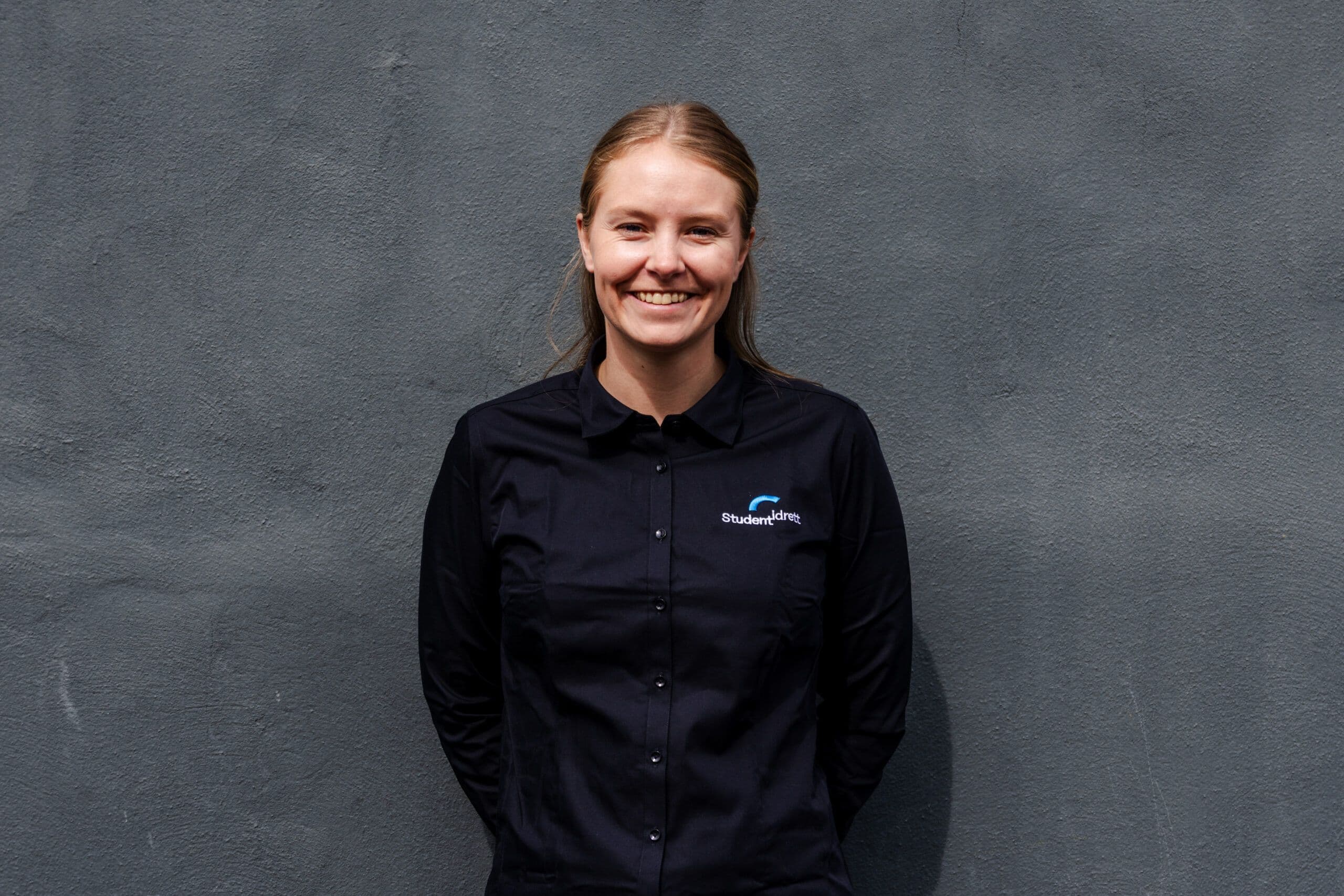 Portrett av en smilende kvinne i svart skjorte med Studentidrett-logo foran en grå betongvegg.