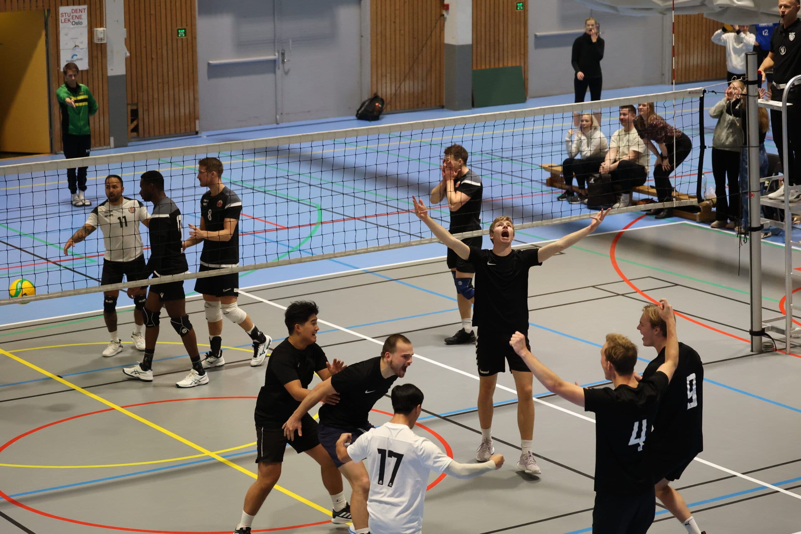 Mannlige volleyballspillere i svarte drakter jubler under en kamp i en idrettshall.