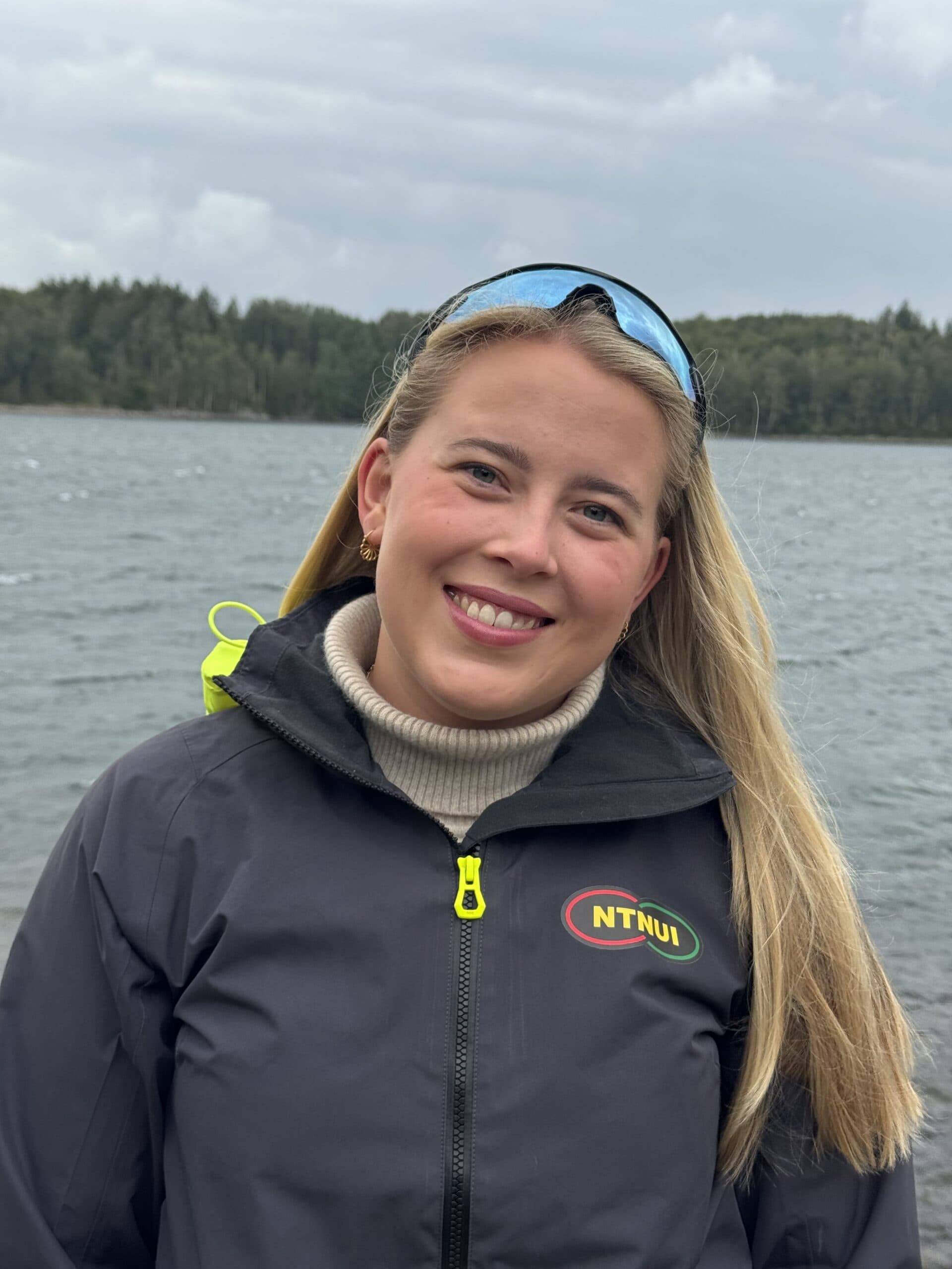 Portrett av en kvinne i en NTNUI-jakke med et vann og skog i bakgrunnen.
