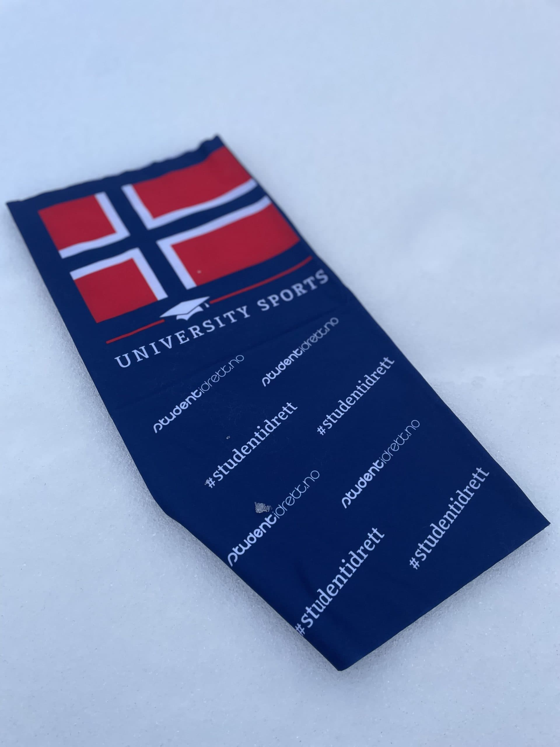 Et blått halsplagg med norsk flagg, teksten University Sports og #studentidrett ligger i snøen.