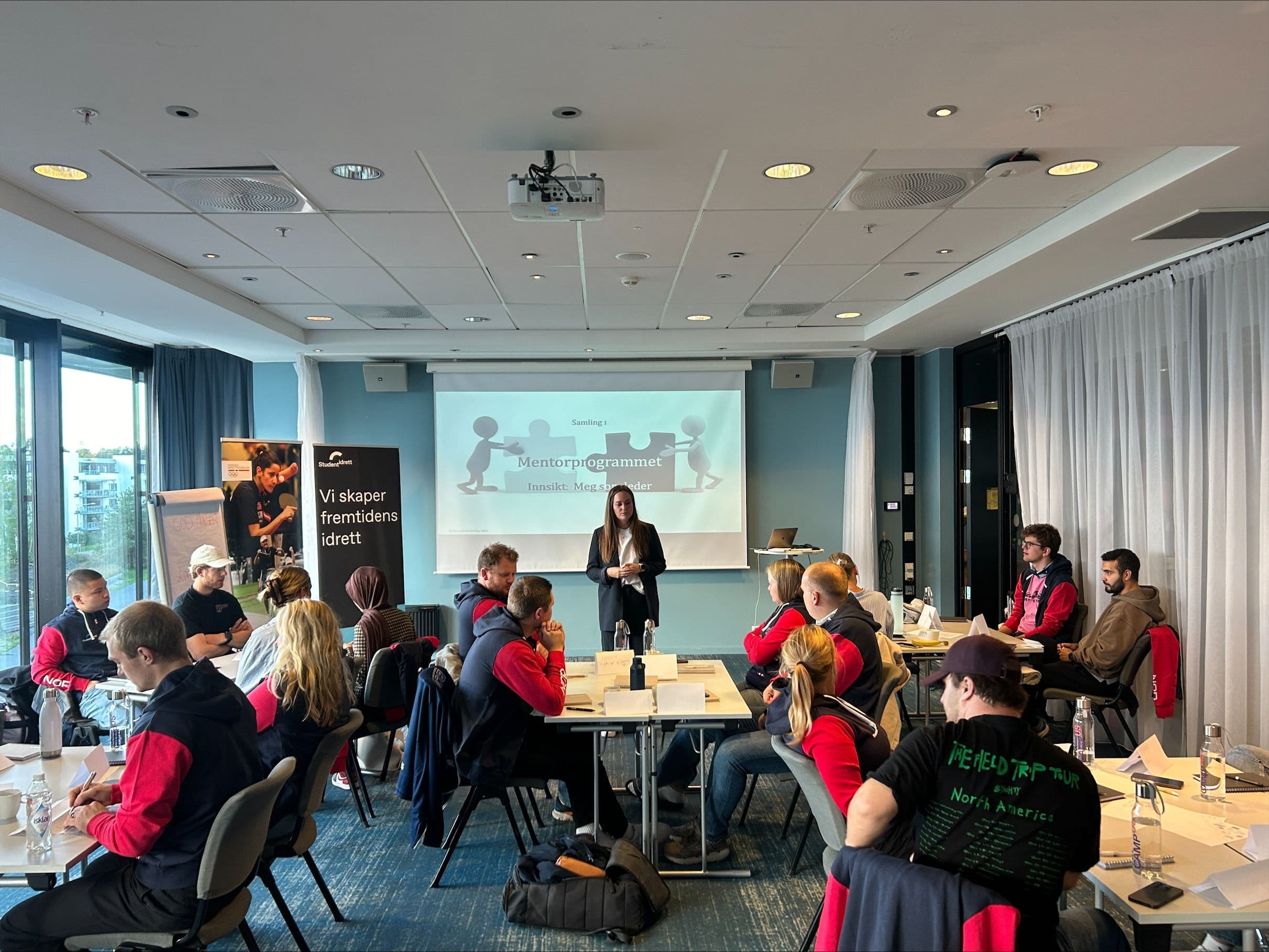 En kvinne holder innlegg foran deltakere ved bord under et seminar i mentorprogrammet.