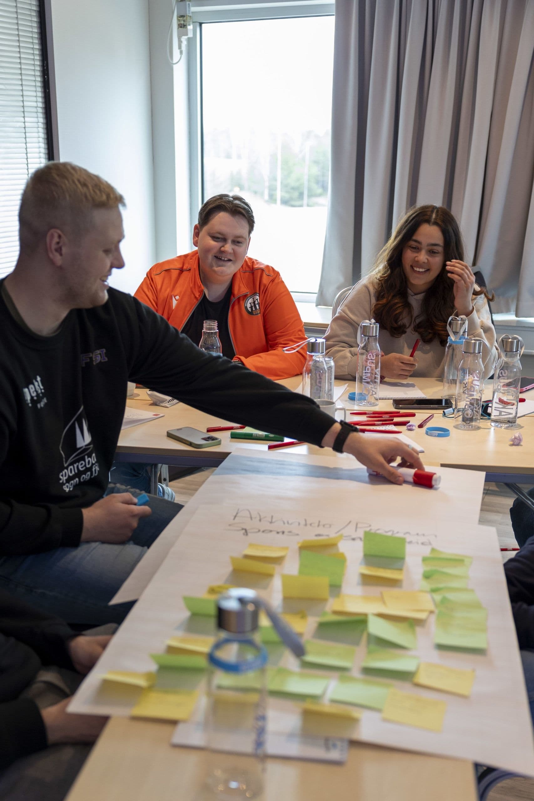 Tre studenter sitter rundt et bord og jobber med gule og grønne post-it-lapper på et stort ark under en workshop.