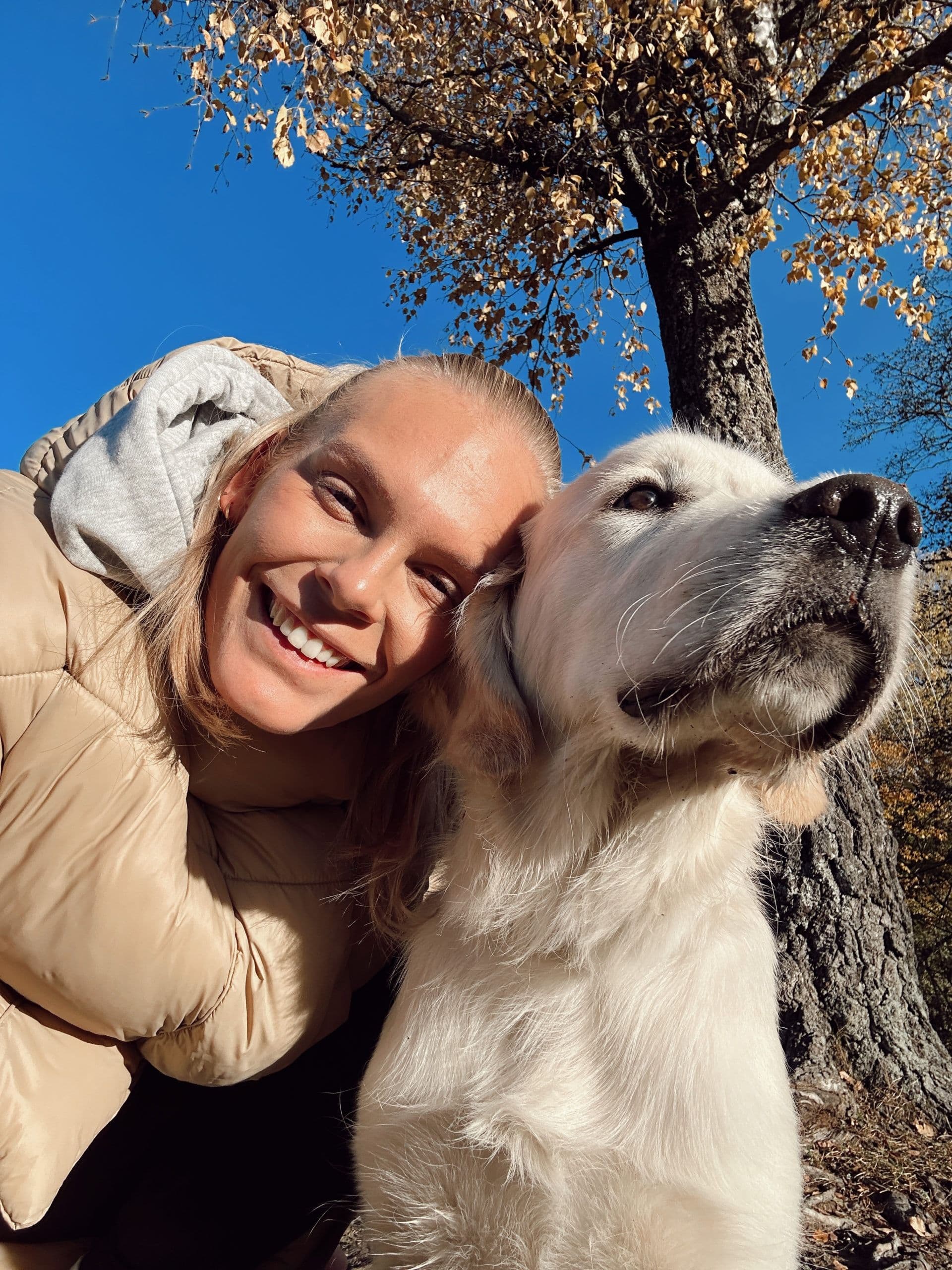 En smilende kvinne i boblejakke lener hodet mot en lys golden retriever ute på en solrik høstdag.